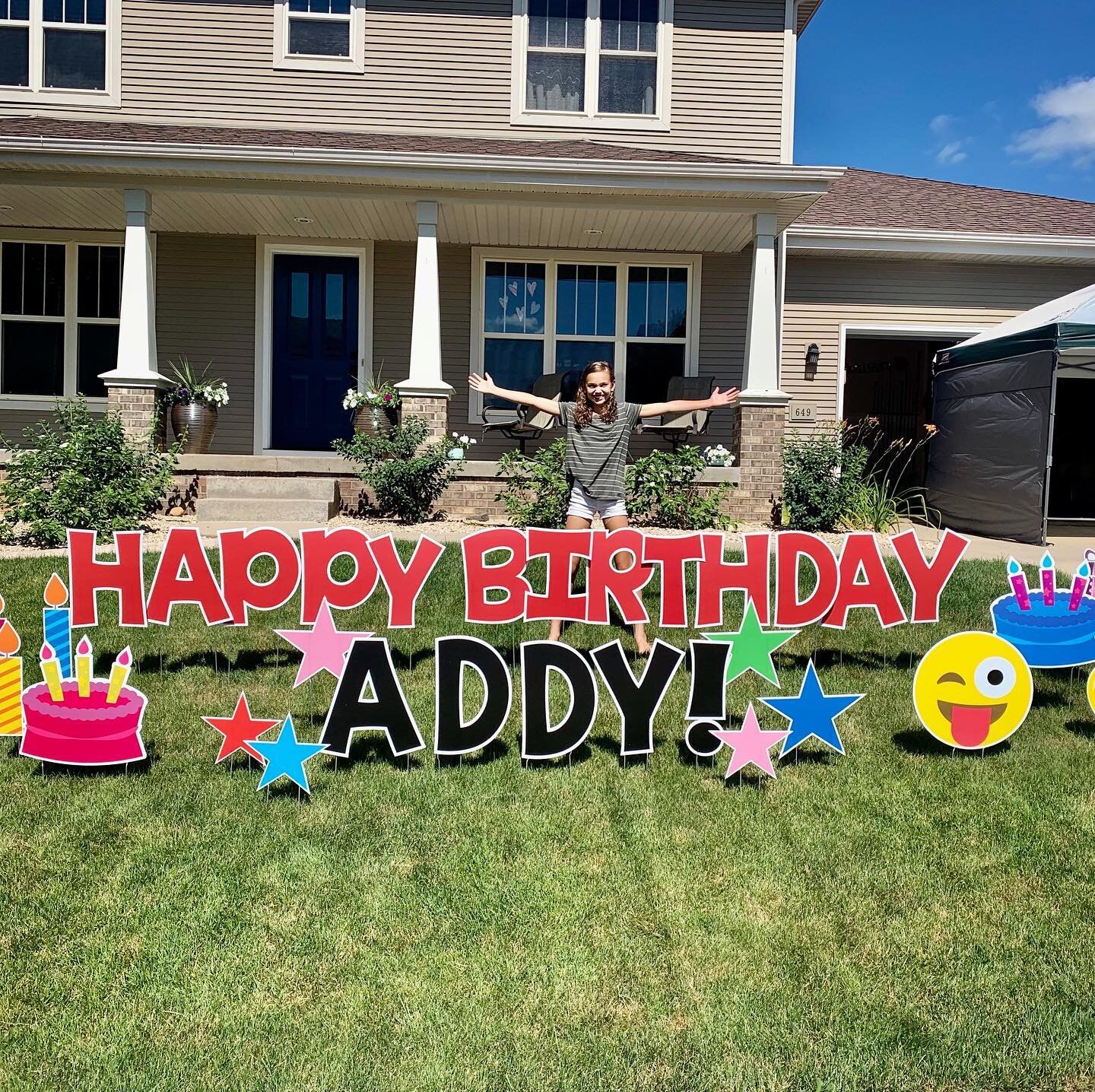 Person standing behind a large sign that says 'Happy Birthday Addy' with colorful decorations on a lawn in front of a house.