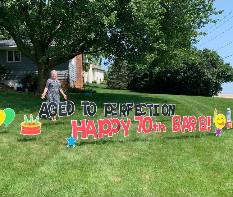 A yard display with large letters spelling 'Aged to Perfection on Happy 70th Barb!' surrounded by colorful decorations including balloons, a cake, and an emoji face. A person stands near the display in a grassy area with trees and a house in the back