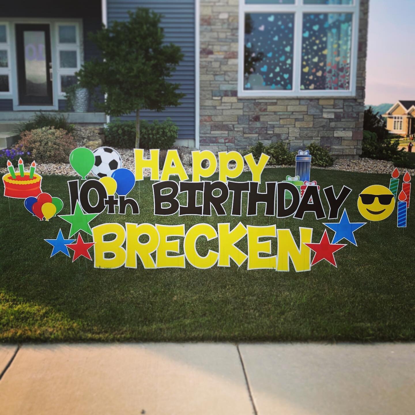 Yard display with colorful letters spelling 'Happy 10th Birthday Brecken' surrounded by balloons, stars, a soccer ball, a cake, emoji, candles, and a gift box.