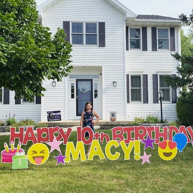 Front yard with a 'Happy 10th Birthday Macy!' sign featuring emojis and balloons in front of a white house, with a young girl standing in front.