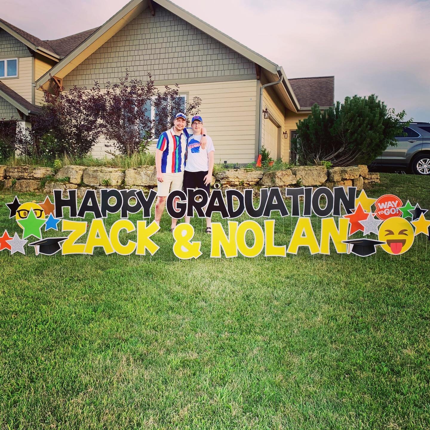Two people standing behind a large outdoor graduation sign with "Happy Graduation Zack & Nolan" in front of a house.