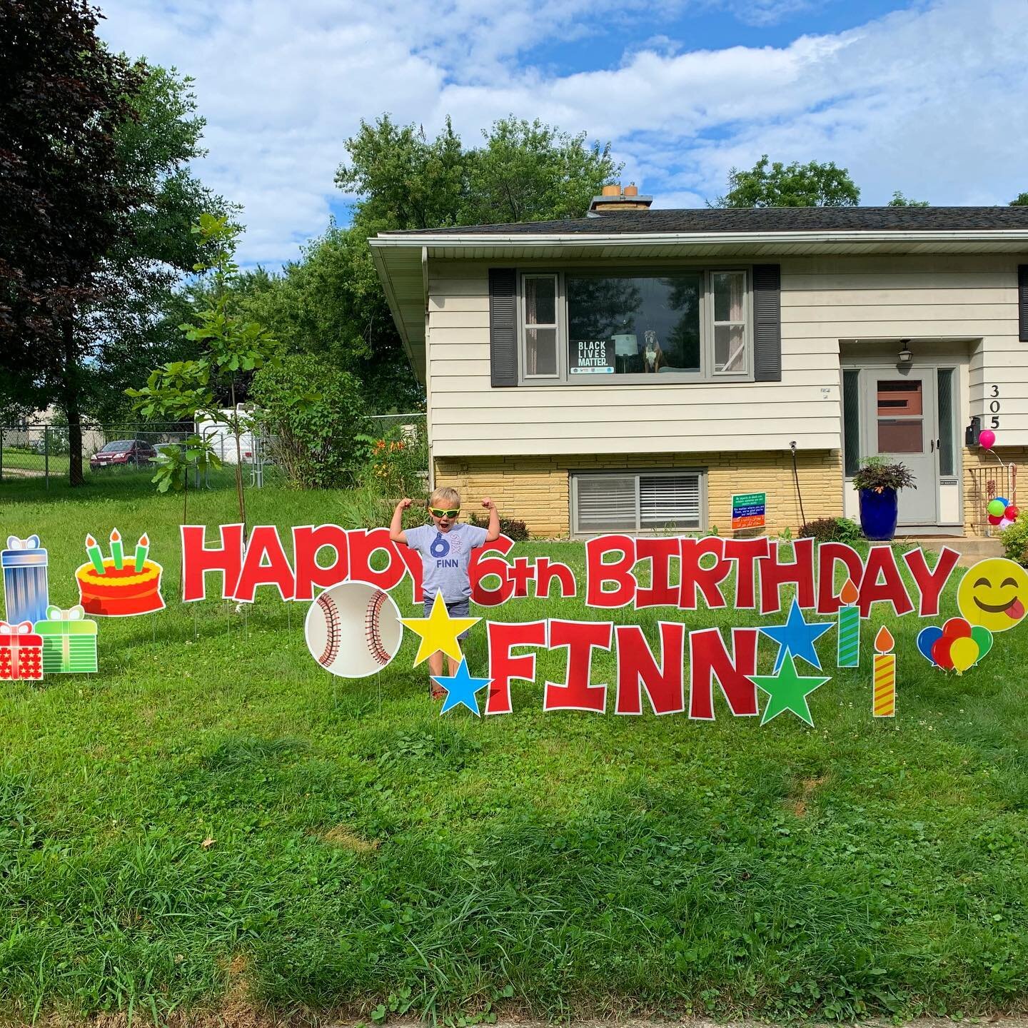 Yard sign saying "Happy 6th Birthday Finn" with cake, baseball, star, and balloon decorations in front of a house. A child is posing next to the sign.