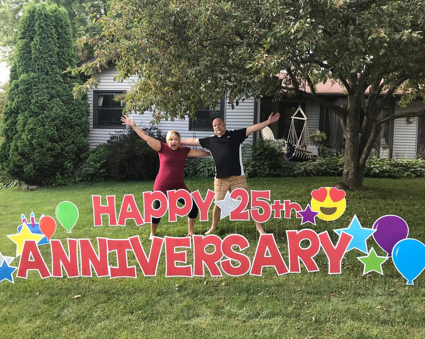 A couple celebrating their 25th anniversary with a large yard sign reading "Happy 25th Anniversary," adorned with balloons and stars, in front of a house with lush greenery.