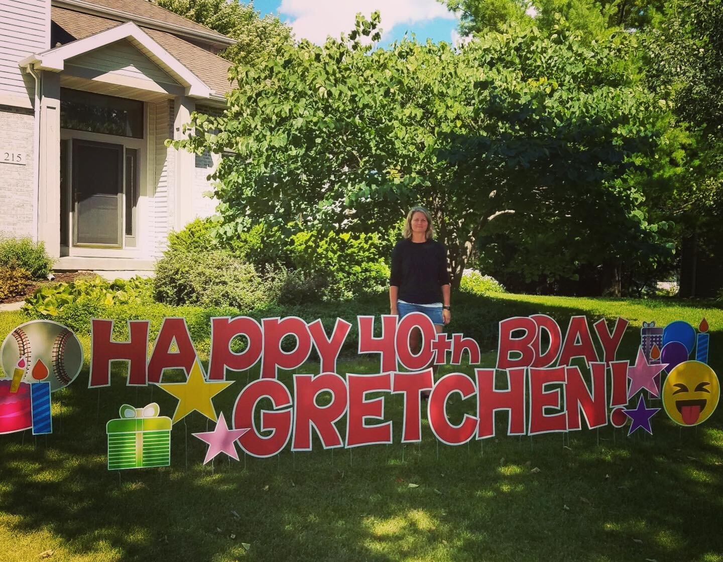 A woman stands behind a large colorful lawn sign in front of a house that reads "Happy 40th Bday Gretchen!" The sign is decorated with images of a baseball, birthday candles, a gift box, stars, and an emoji. There are trees and shrubs in the backgrou