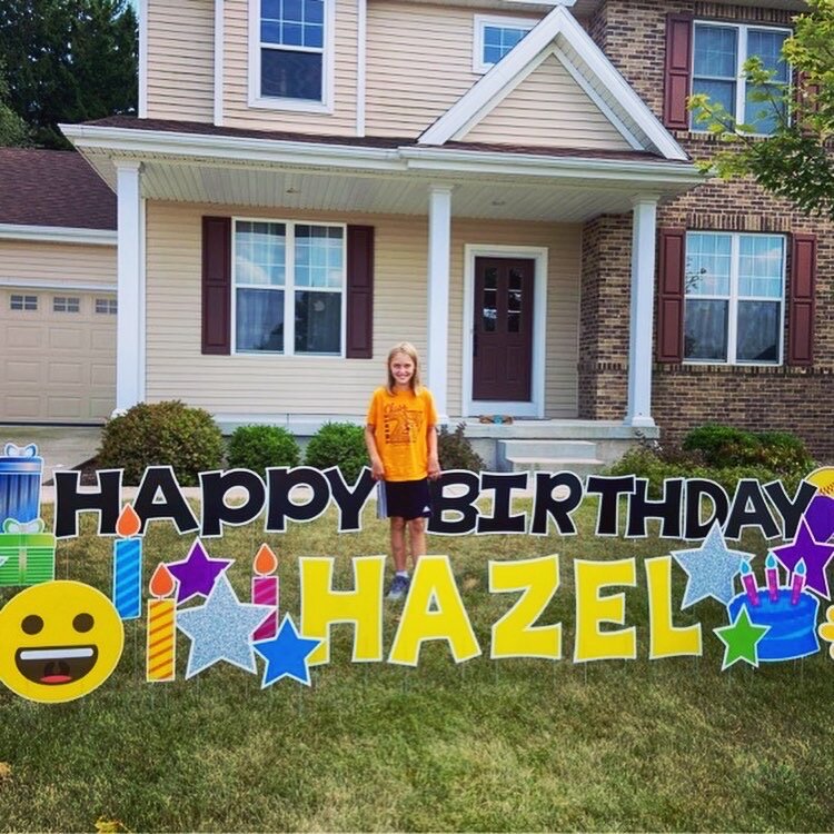 Child standing in front of house with large outdoor birthday sign reading 'Happy Birthday Hazel,' decorated with colorful stars and smiley faces.