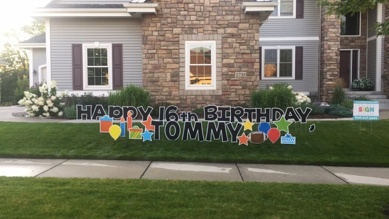 Front yard of a house with a large sign displaying "Happy 16th Birthday Tommy" along with colorful decorations like gifts and balloons.