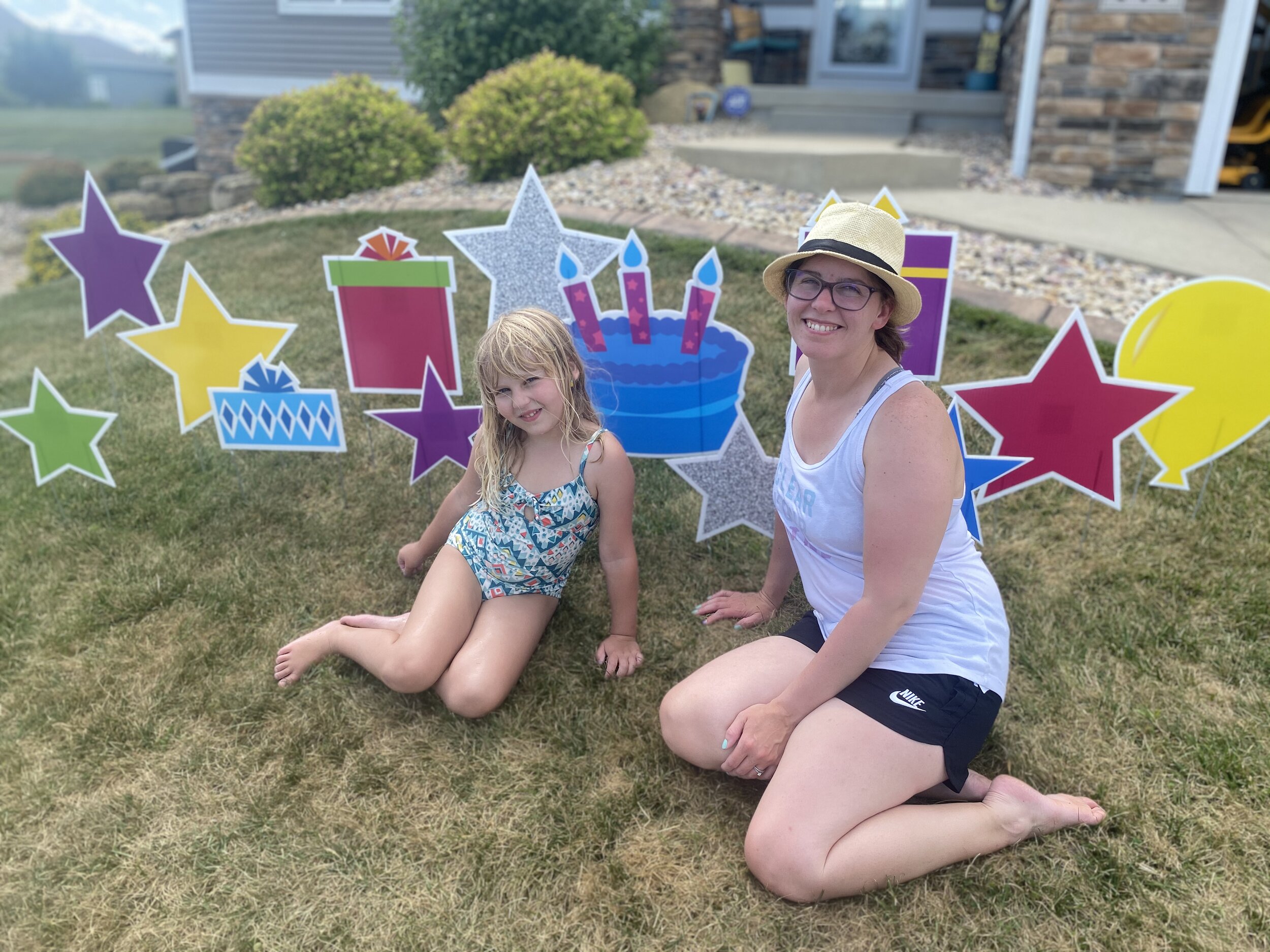A girl in a floral swimsuit and a woman in a hat and tank top are sitting on grass, surrounded by colorful decorations including stars, presents, and a birthday cake cutout with candles. They are outdoors, in front of a house.
