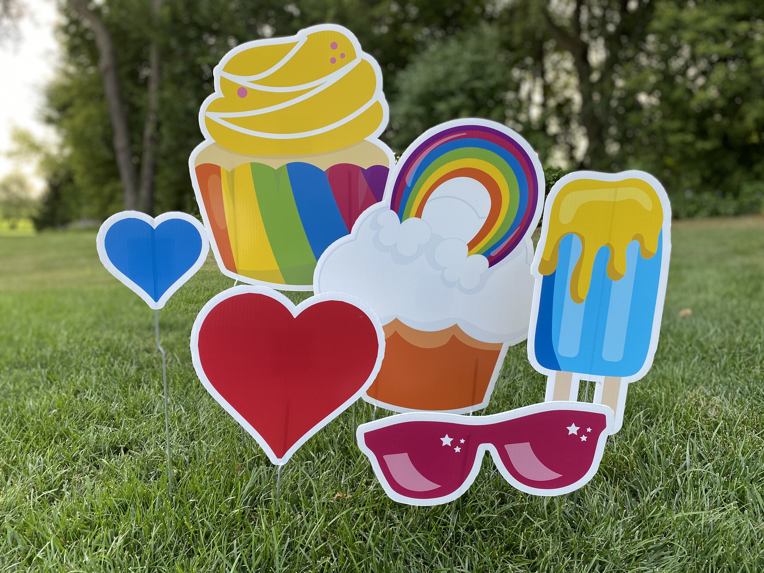 Colorful yard signs on grass including a rainbow cupcake, blue heart, red heart, sunglasses, popsicle, and rainbow.