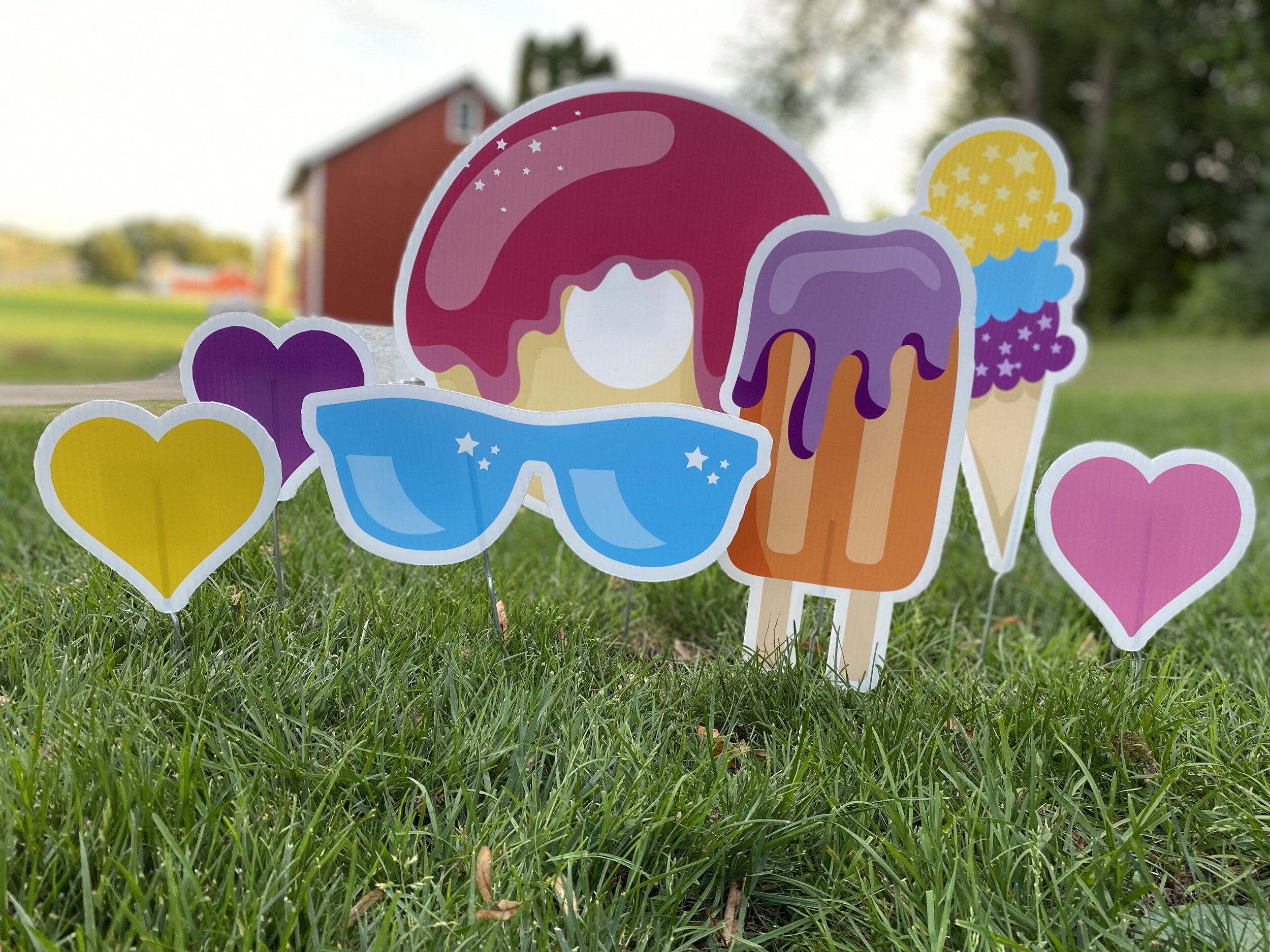 Colorful yard signs featuring a donut, sunglasses, a popsicle, an ice cream cone, and heart shapes on green grass with a red barn in the background.