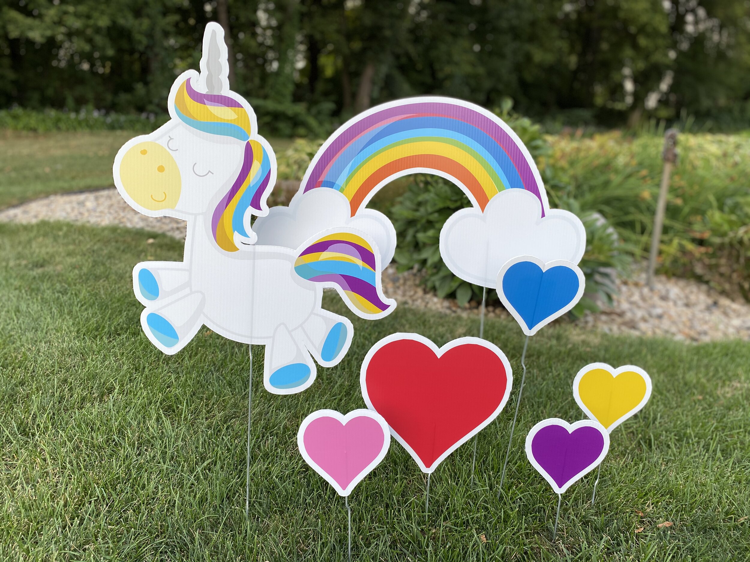Garden decorations featuring a colorful cartoon unicorn with a rainbow mane and tail, set against a rainbow with fluffy clouds. Surrounding the unicorn are colorful heart-shaped cutouts on a grassy lawn.