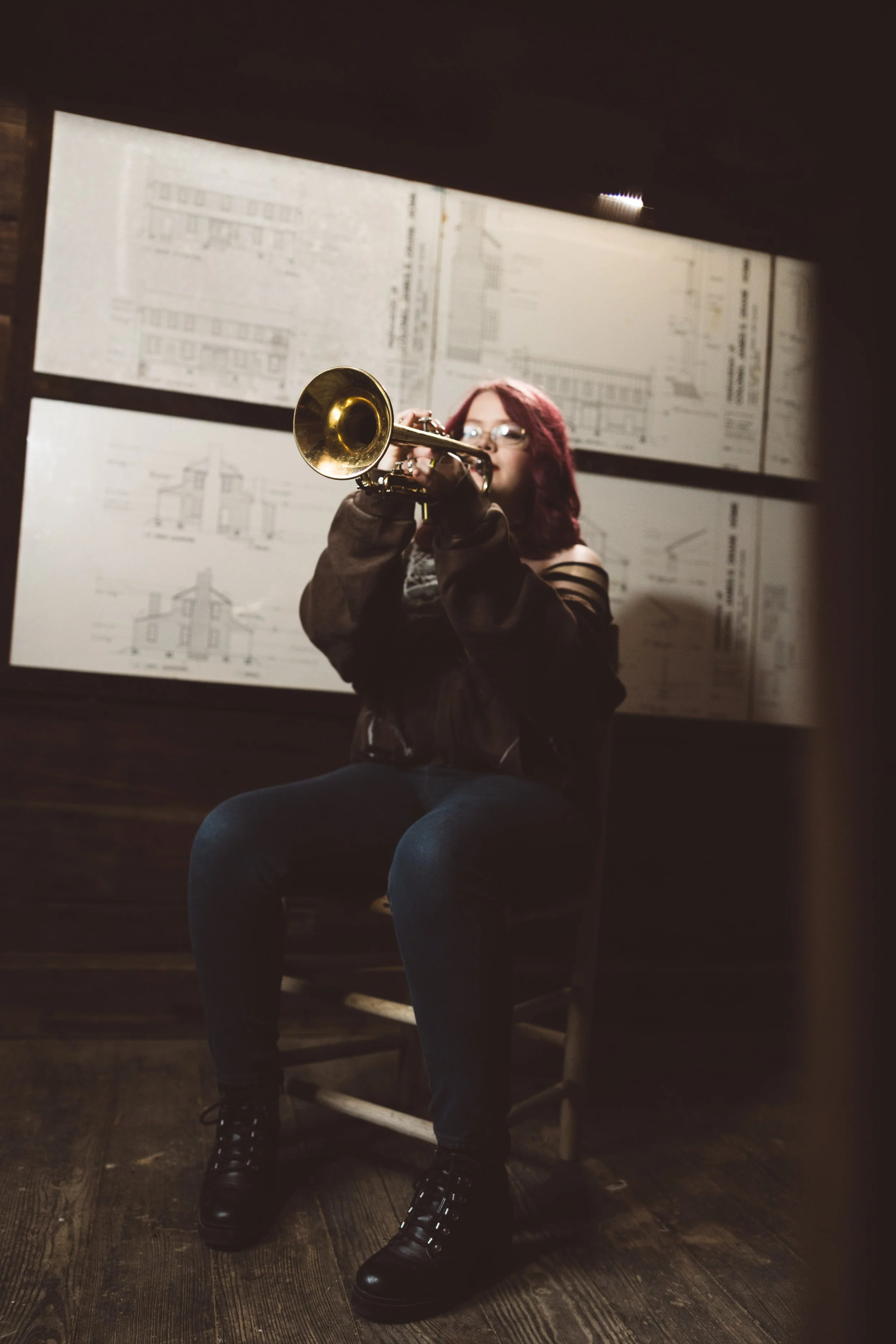 A woman with red hair and glasses is sitting on a chair, playing a trumpet, with architectural drawings on the wall behind her.