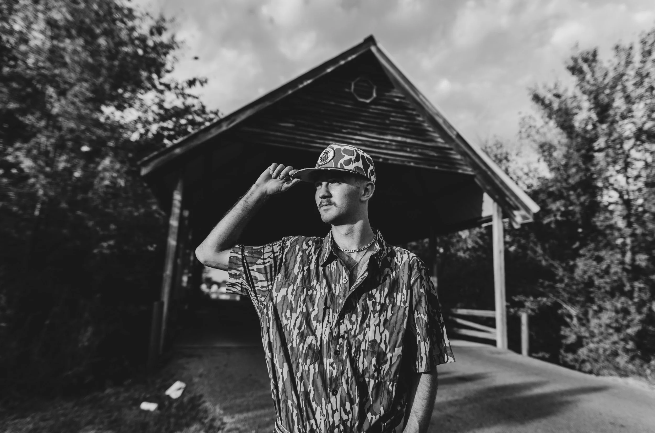A young man standing outside near a wooden building with trees in the background, wearing a camouflage patterned shirt and a colorful camouflage hat, holding the brim of his hat with one hand, in black and white photo.