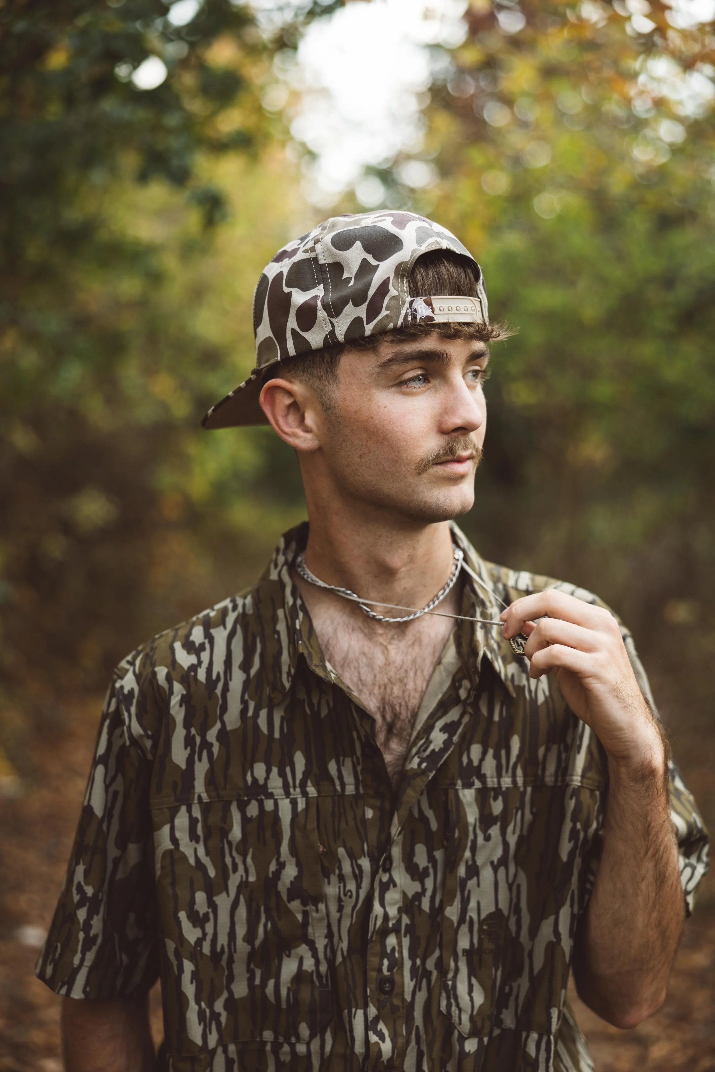 A young man with a patterned camo hat and shirt, holding a chain necklace, standing outdoors with a blurred background of trees and foliage.