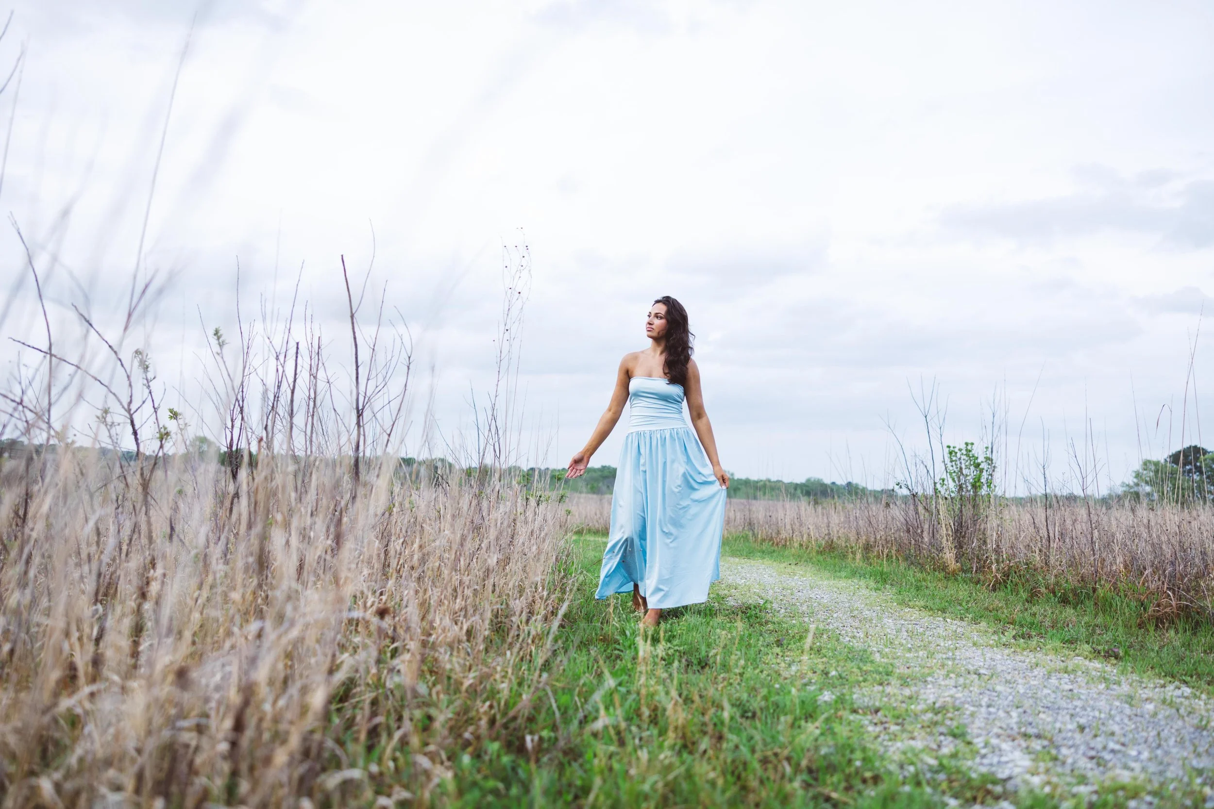 A woman walking through a grassy field on a dirt path, wearing a long, strapless blue dress, with tall grass on both sides and an overcast sky above.