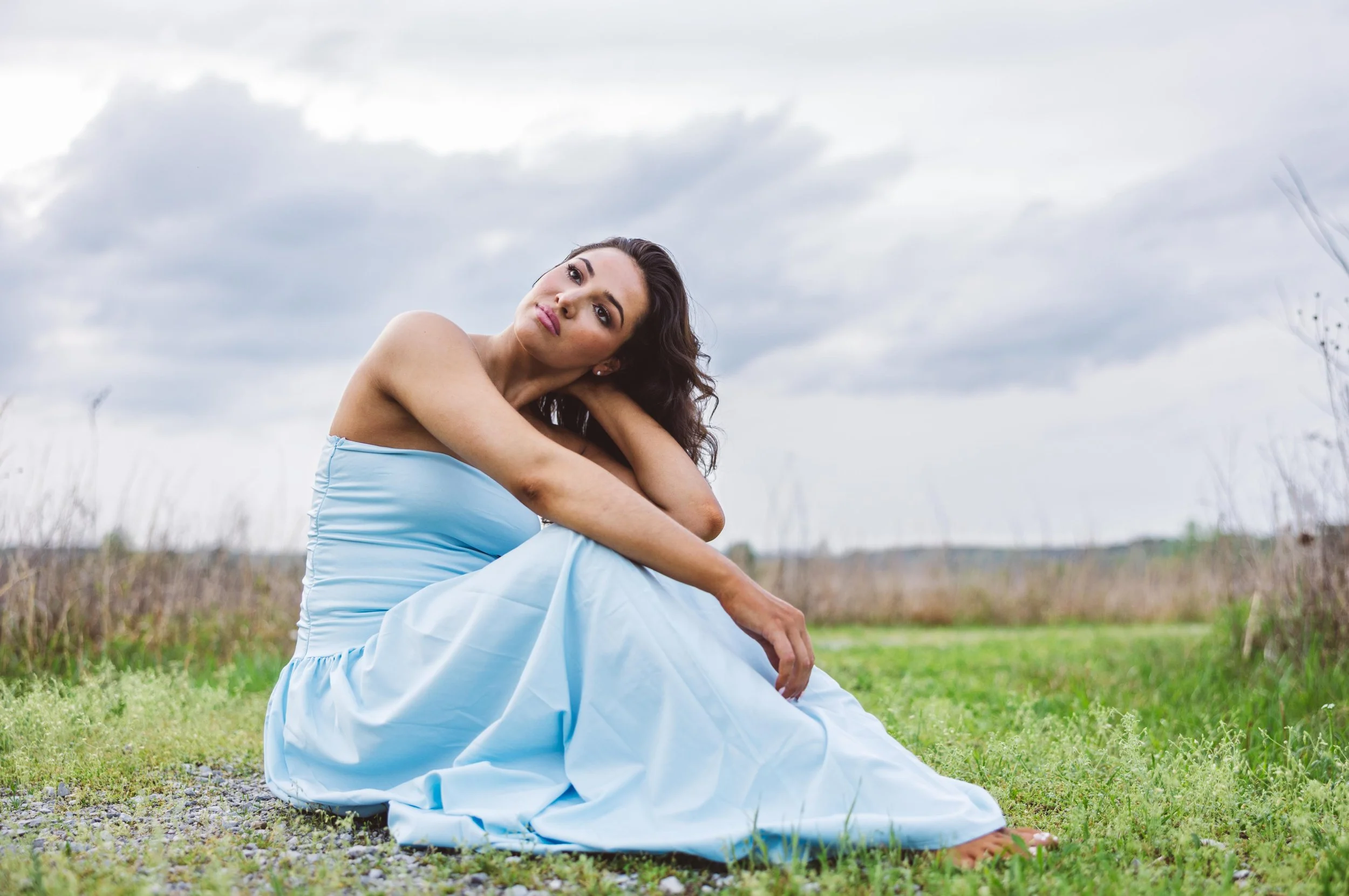 A woman in a light blue strapless dress sitting on grass in a field with cloudy sky.