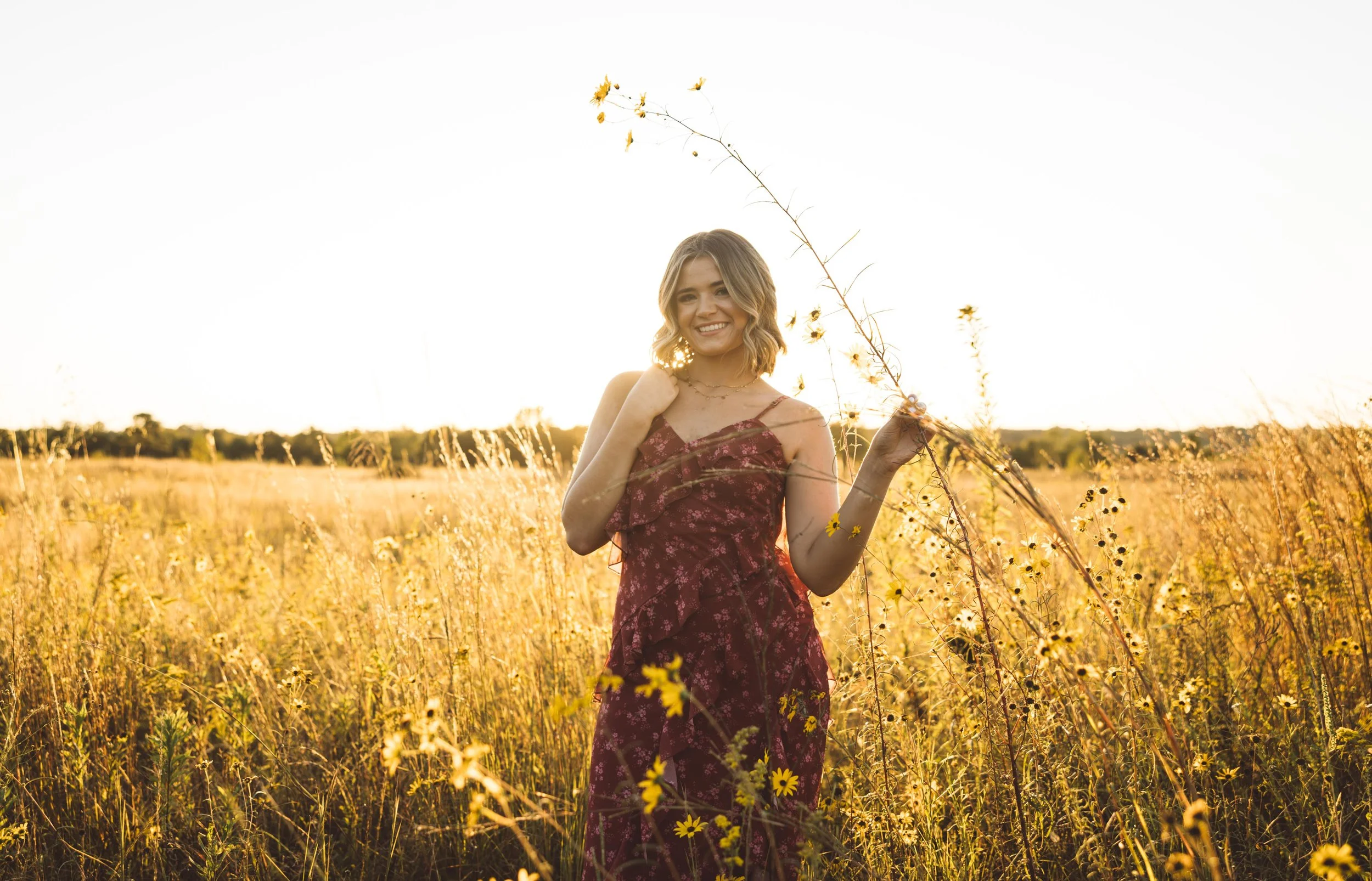 A young woman with blonde hair smiling in a yellow flowered field at sunset, wearing a maroon floral dress.