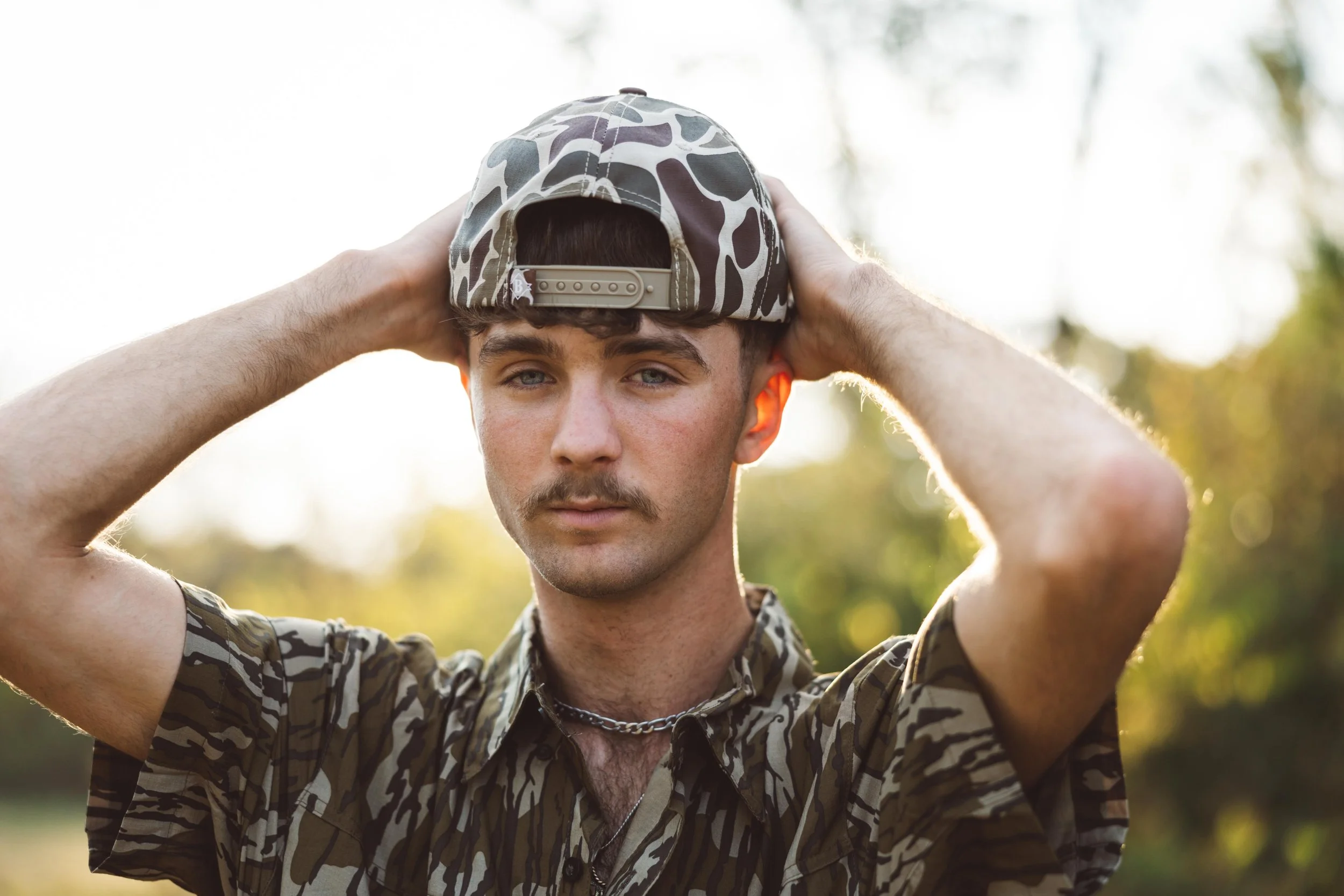 A young man wearing a camo patterned cap and shirt, with a chain necklace, stands outdoors in natural light, adjusting his cap with both hands, with blurred trees in the background.
