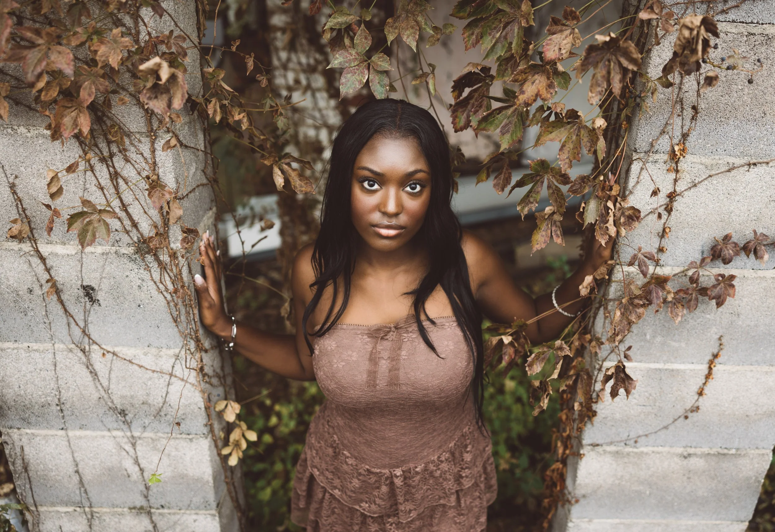 A woman in a brown lace dress is looking directly at the camera while holding onto stone walls covered with vines.