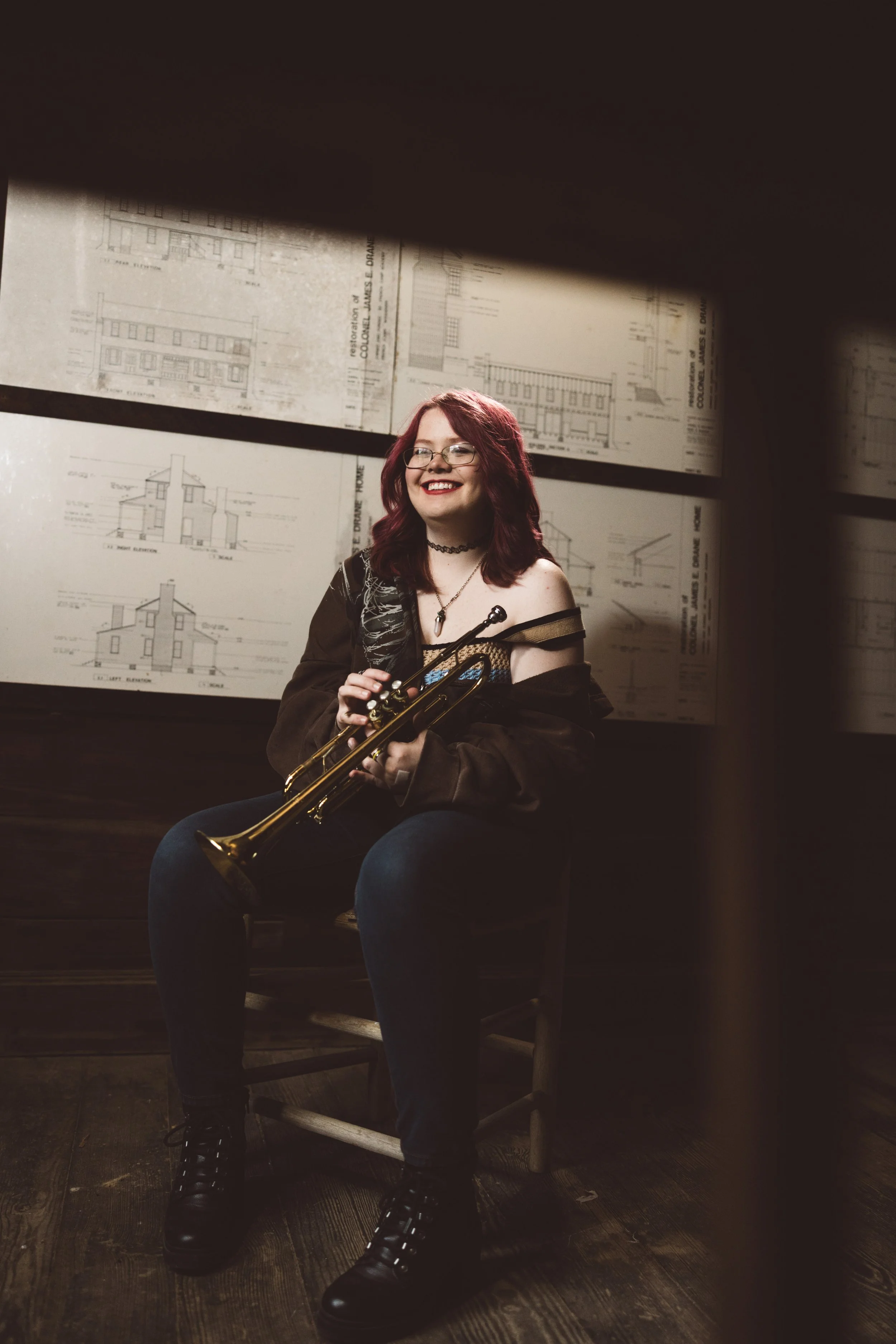 Young woman with red hair, glasses, and a choker necklace sitting on a wooden chair holding a brass trumpet, smiling at the camera. She is in a room with architectural blueprints on the wall behind her.