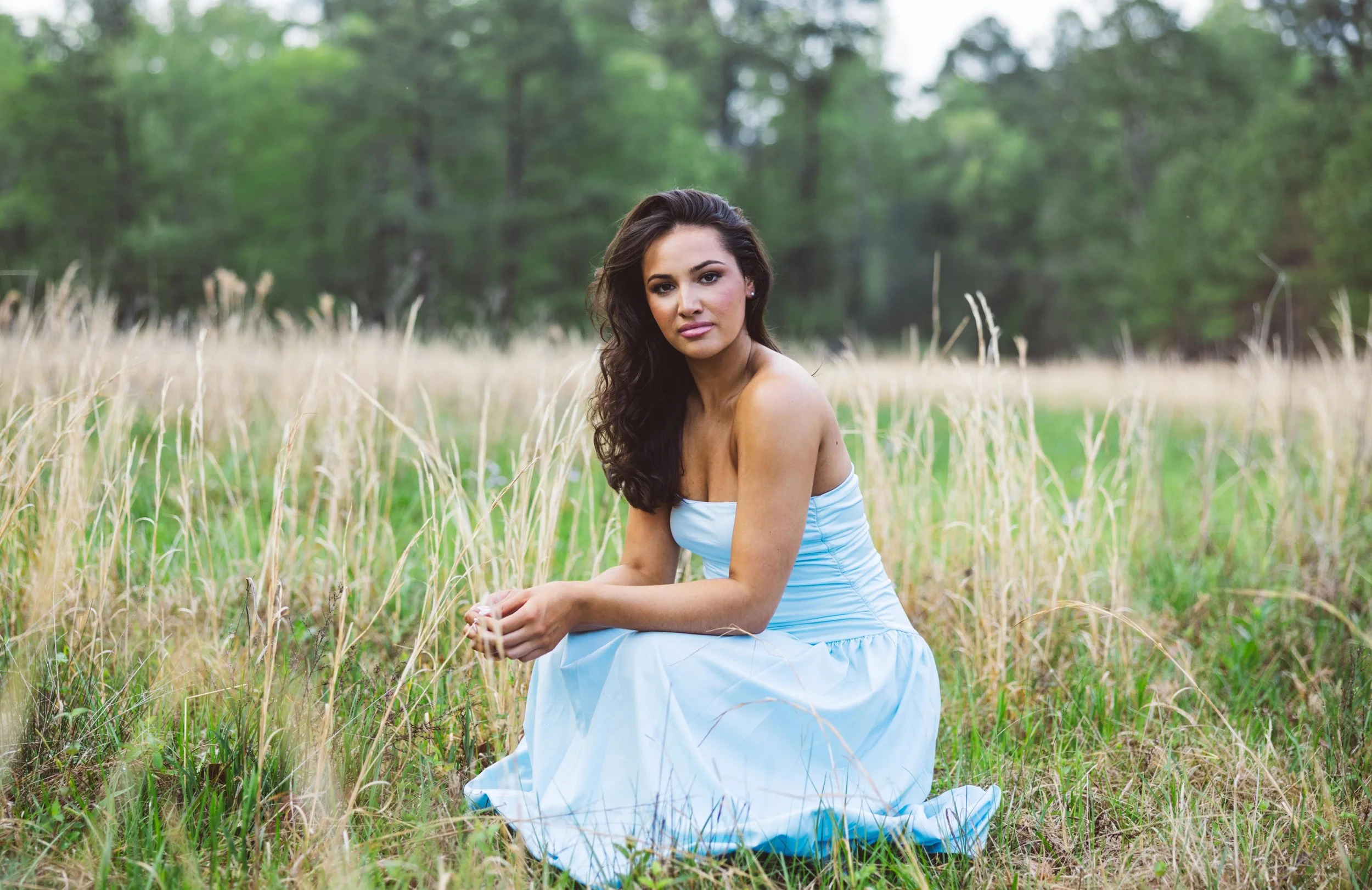 A woman with dark, wavy hair seated on grass in a field of tall grass and wildflowers, with trees in the background, wearing a strapless light blue dress.