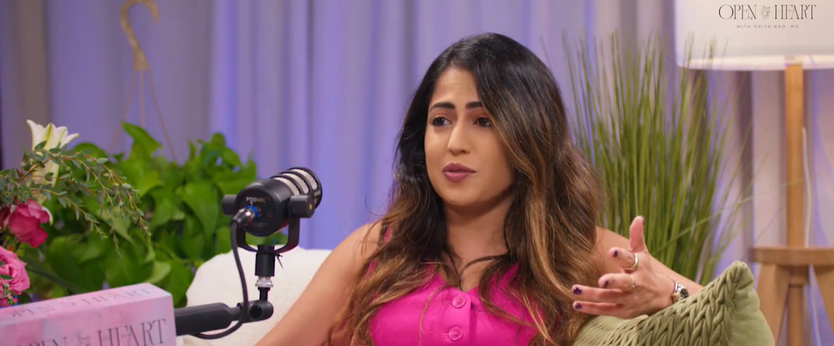 A woman with long, wavy hair wearing a pink top speaking into a microphone during a recorded interview or podcast. She is gesturing with her right hand, seated on a comfortable chair with a green pillow, with floral and indoor plants in the background.