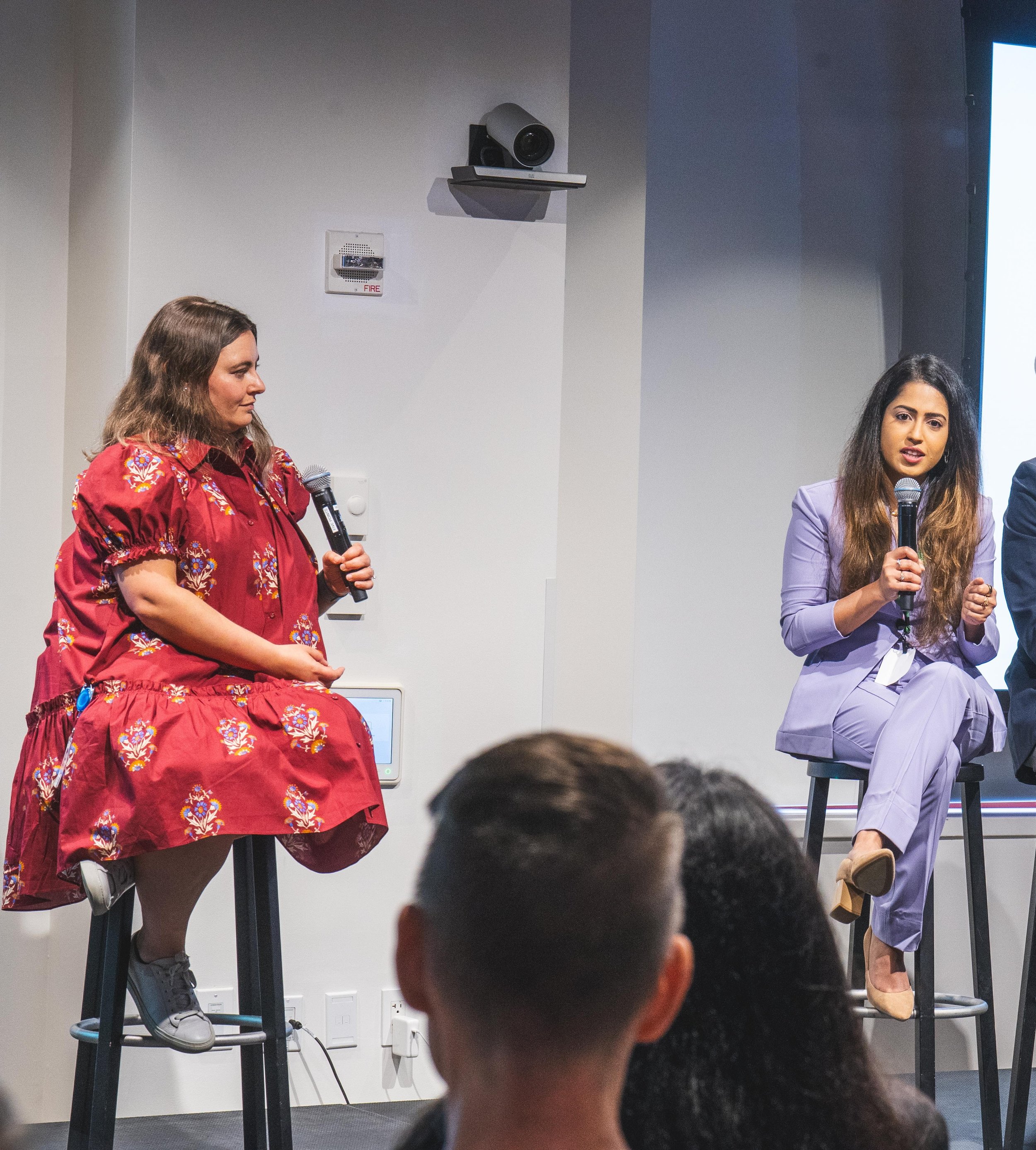 Two women sitting on stools having a discussion, each holding a microphone, on a stage with audience members in the foreground.