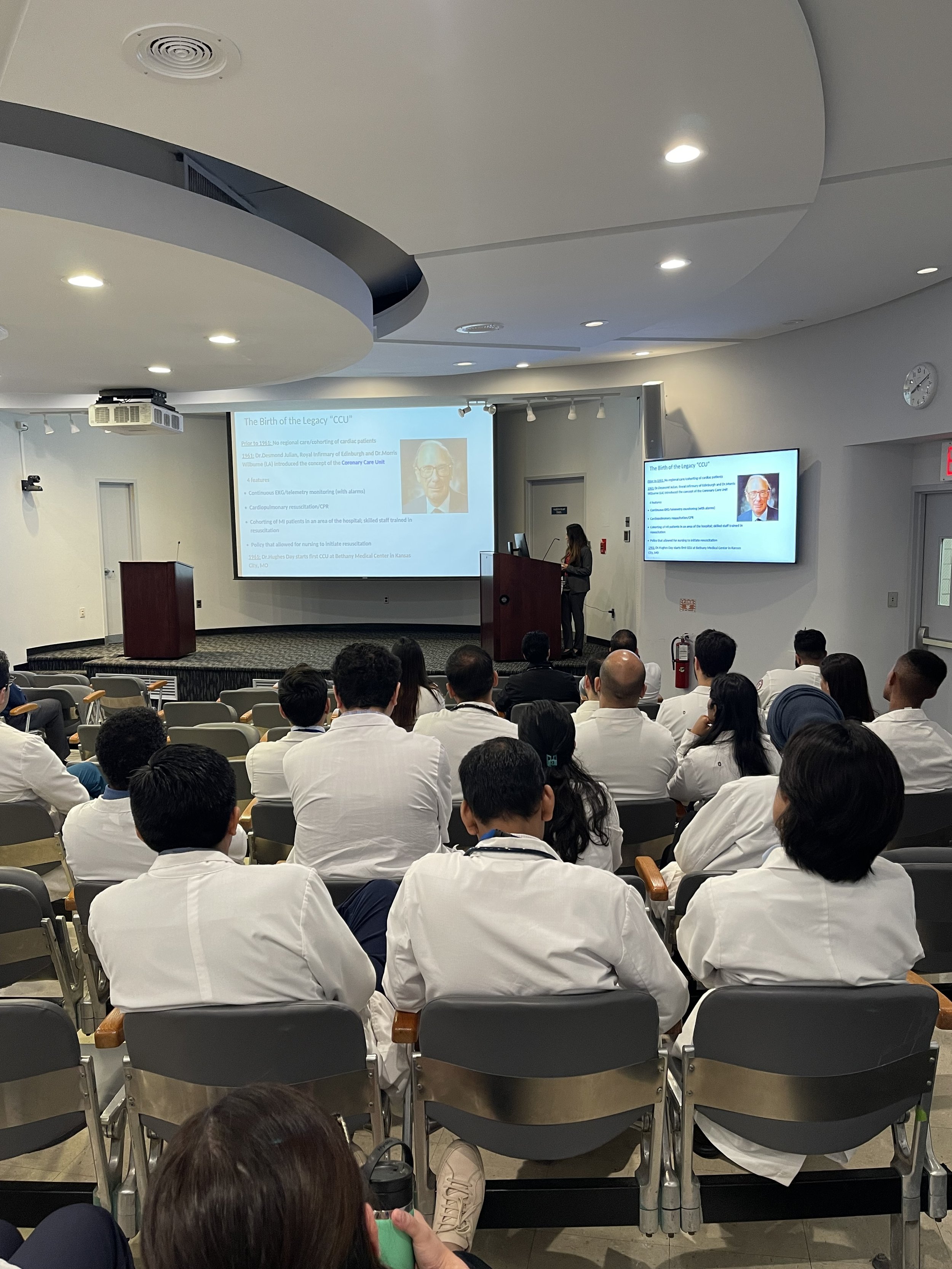 Rosy Thachil, MD, MBA, FACC along with healthcare professionals in white coats seated and listening to a speaker at a podium. The presentation slide discusses the history of the CCU and features a portrait of a man.