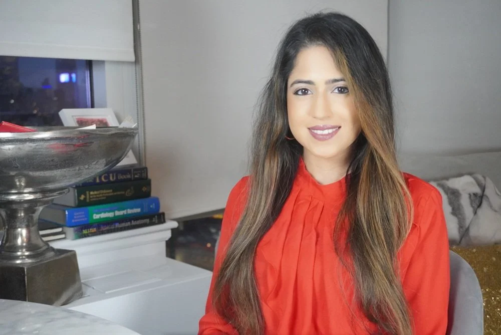 A woman with long brown hair, wearing a red blouse, sitting indoors in front of a gray wall, smiling at the camera.