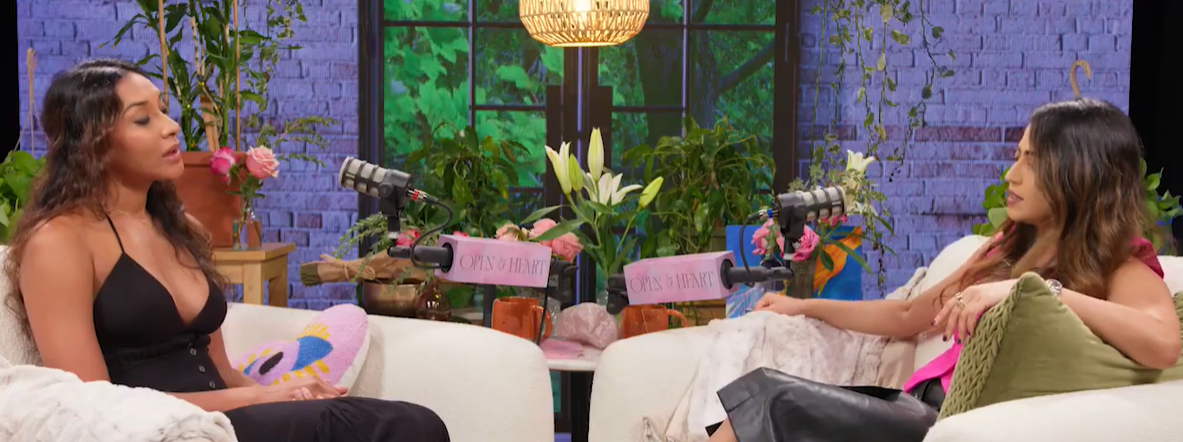 Two women having a conversation in a cozy, plant-filled studio setting with microphones, flowers, and a decorative brick backdrop.