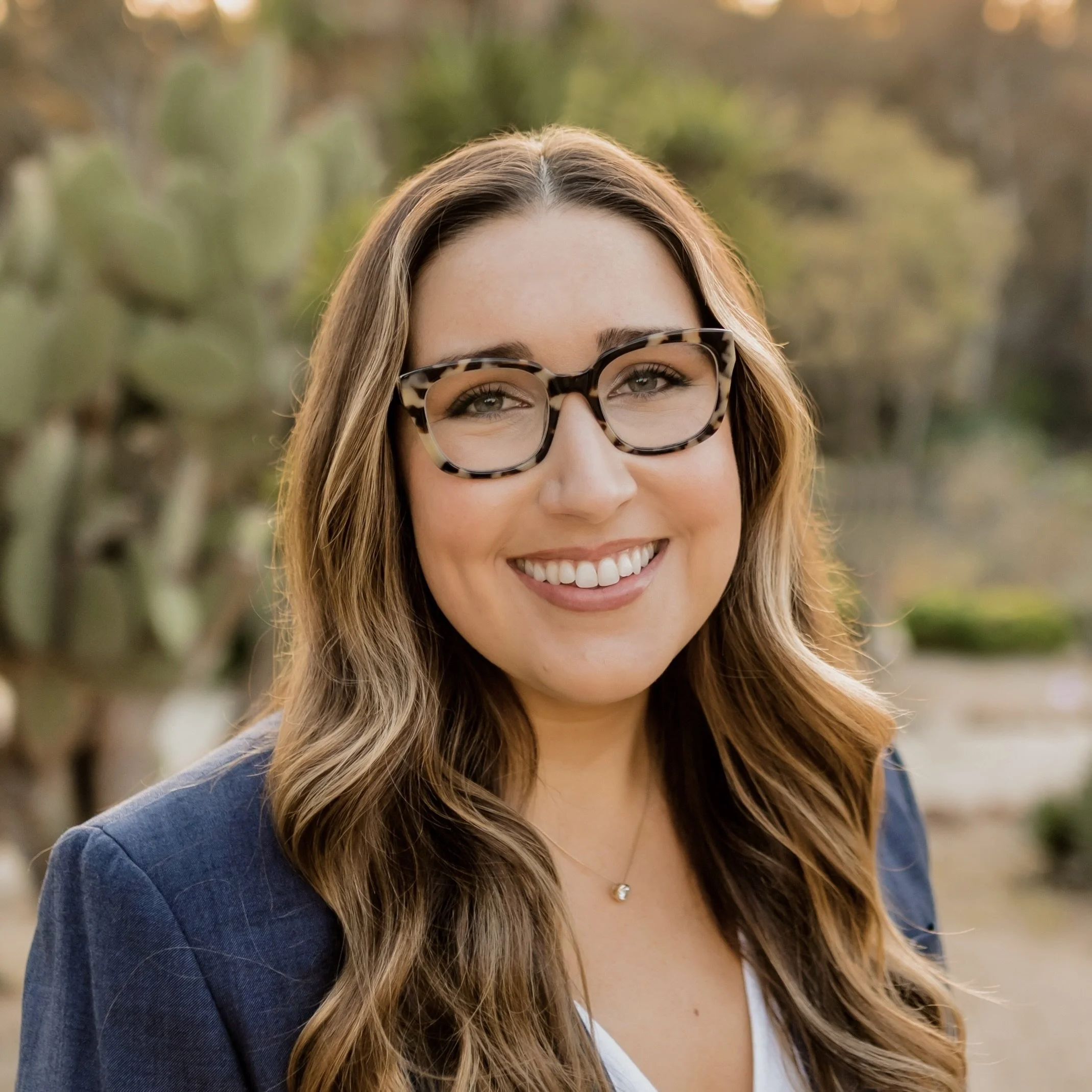 A woman with long wavy brown hair wearing glasses and a navy blazer and white blouse, smiling against a nature background.
