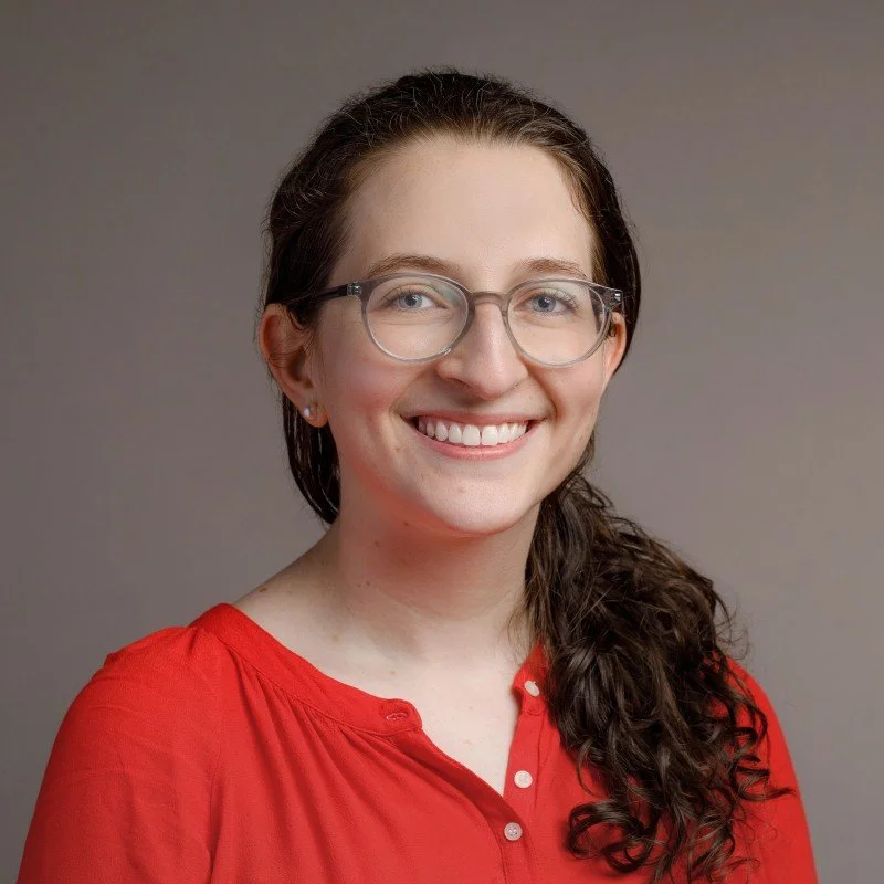 Sara Winograd, PsD, A woman with long curly brown hair wearing glasses and a red blouse, smiling against a plain gray background