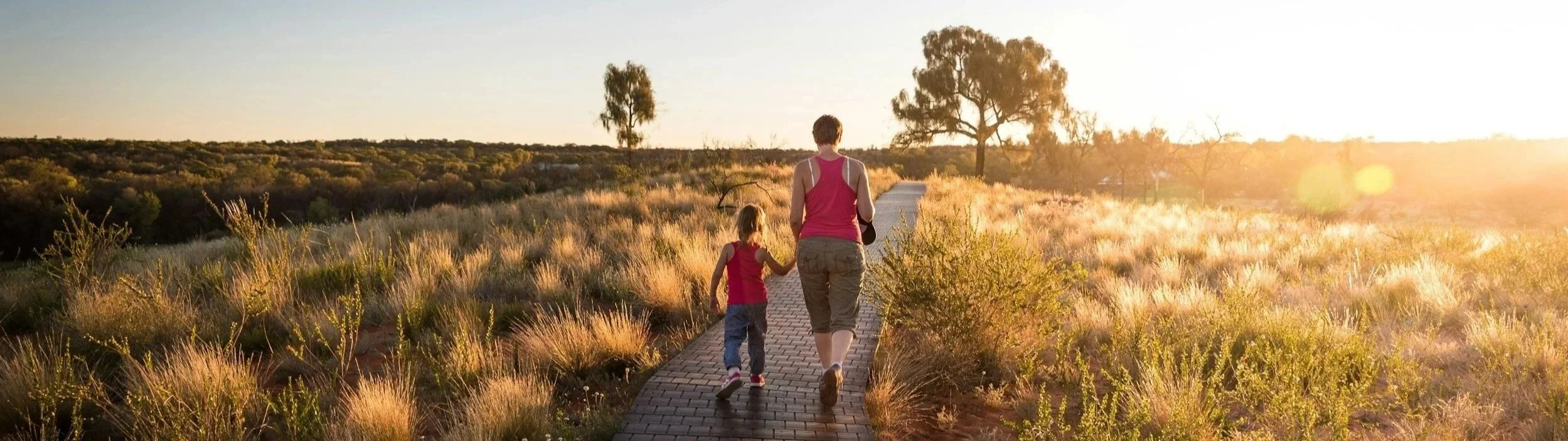 A woman and a young girl walk hand in hand on a brick pathway through a dry, grassy landscape at sunset, with trees in the distance.