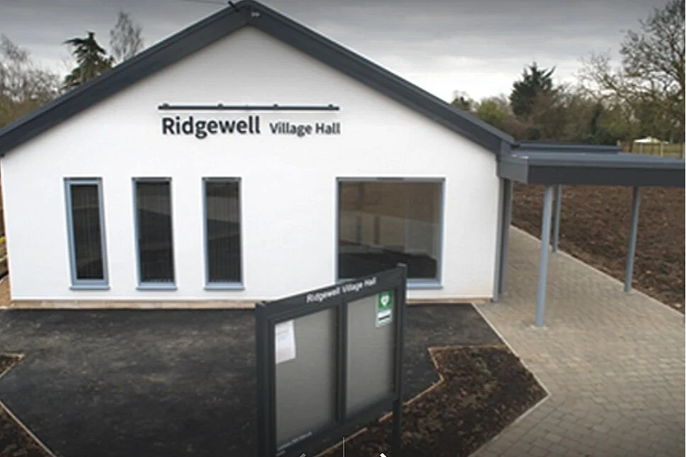 A new white village hall building with the sign 'Ridgewell Village Hall' on the front, featuring four tall narrow windows and a covered walkway.