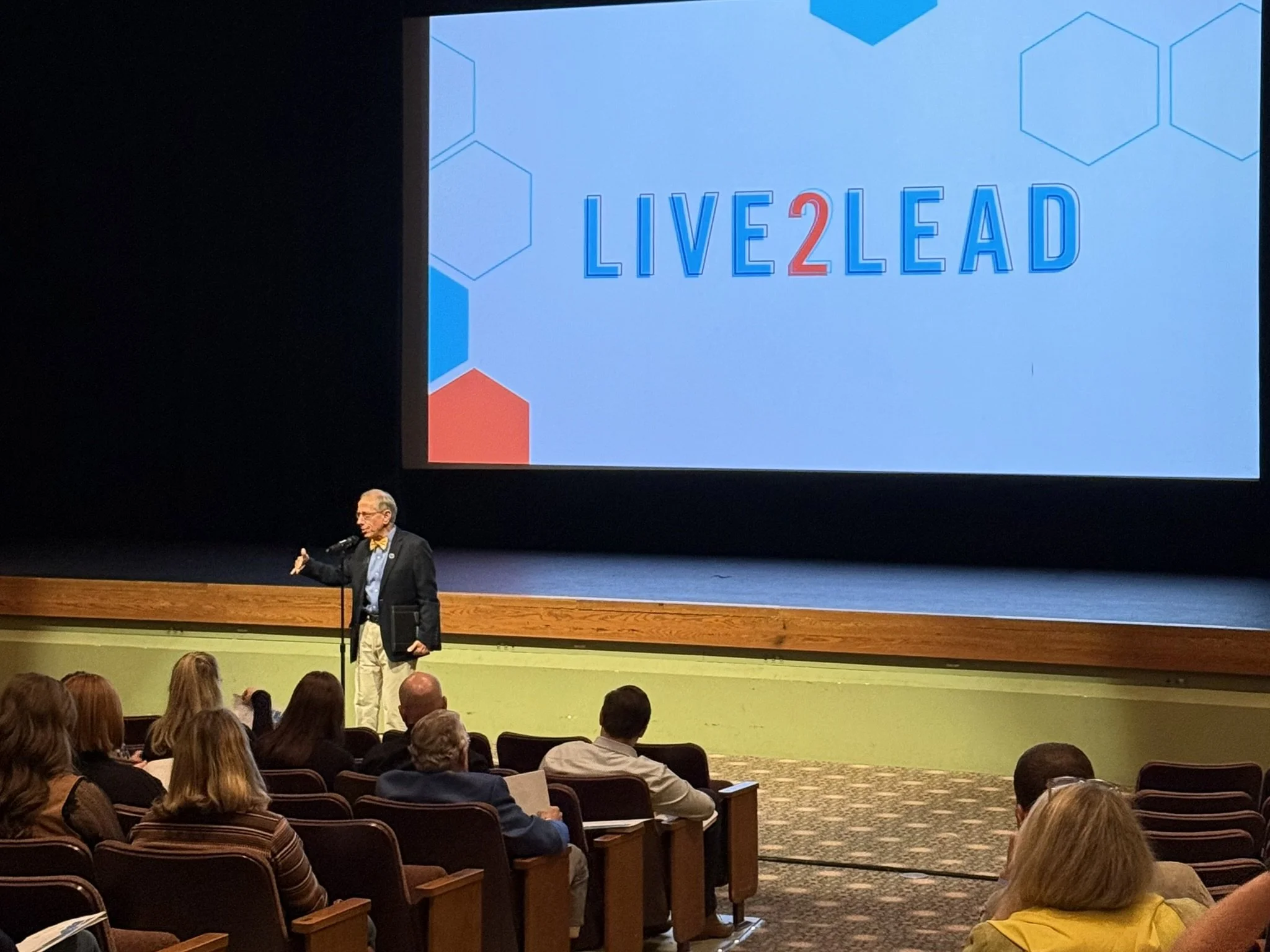 A man with gray hair and glasses, dressed in a suit with a yellow bow tie, giving a presentation on a stage in front of an audience. Behind him, a large screen displays the words "LIVE 2 LEAD" with a blue and red color theme and geometric graphics.