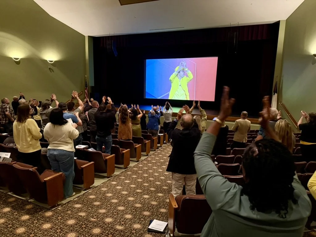 Audience members in a theater clapping and raising their hands while watching a large video screen showing a person in a yellow jacket with hands near their face.