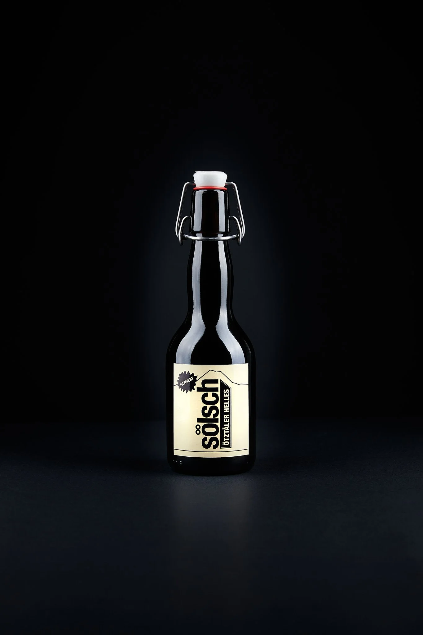 Eine schwarze Flasche mit weißem Etikett und einem Korkverschluss, auf schwarzem Hintergrund. Fotografiert von Sarah Neuner.