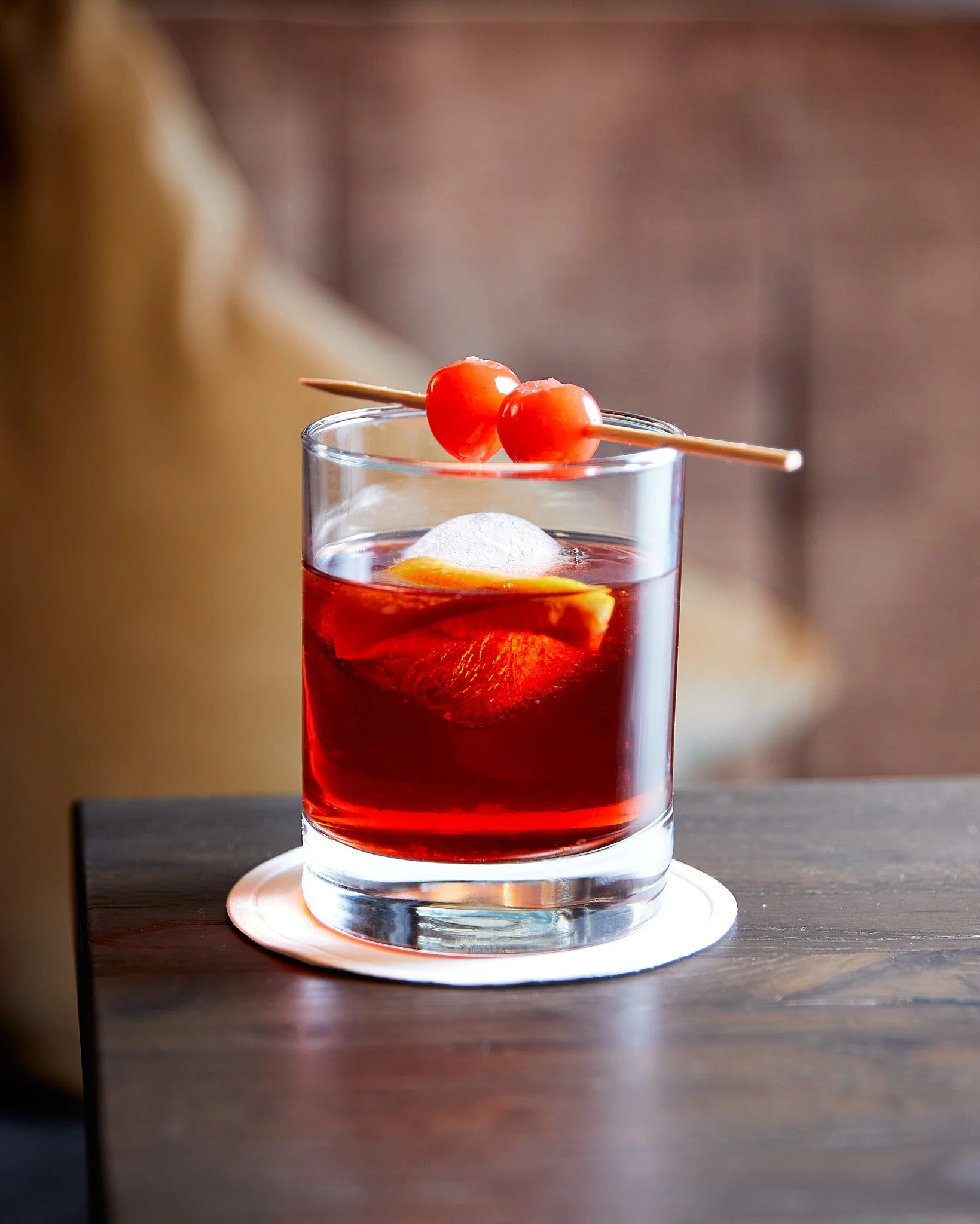 Ein Glas Rotwein mit Eis im Glas, garniert mit einer Zitronenscheibe und einem Cocktailspieß mit zwei Kirschen, auf einem runden weißen Untersetzer auf einem dunklen Holztisch. Fotografiert von Sarah Neuner.