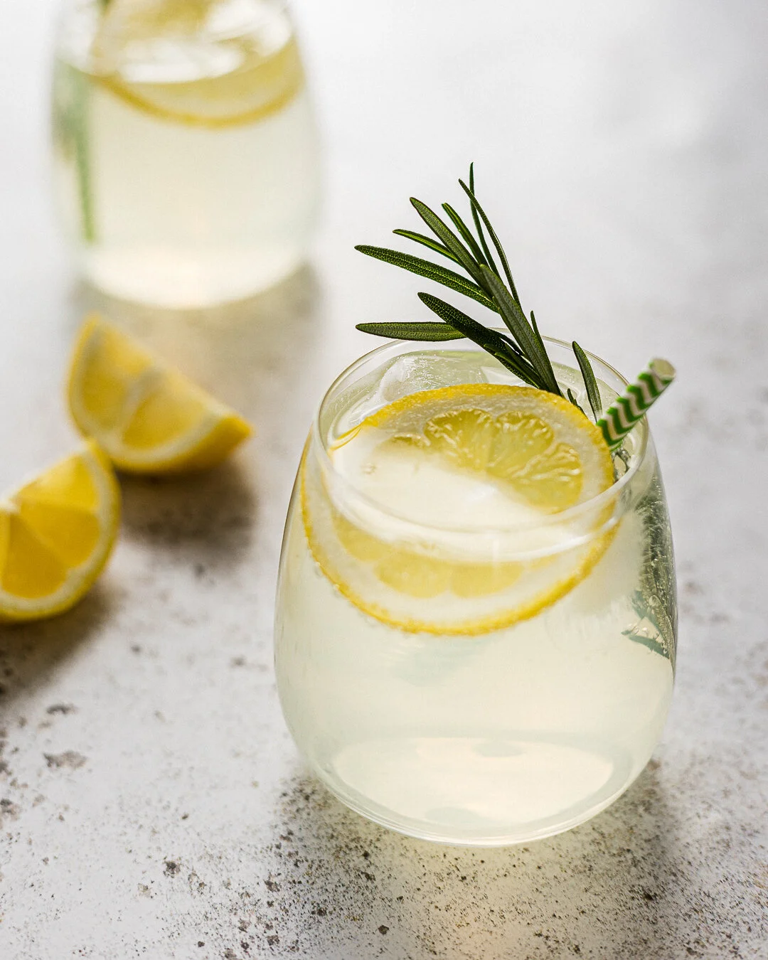 Ein Glas Zitronenwasser mit Zitronenscheiben, Rosmarinzweigen und Trinkhalm, daneben mehr Zitronen auf einem hellen Hintergrund. Fotografiert von Sarah Neuner.