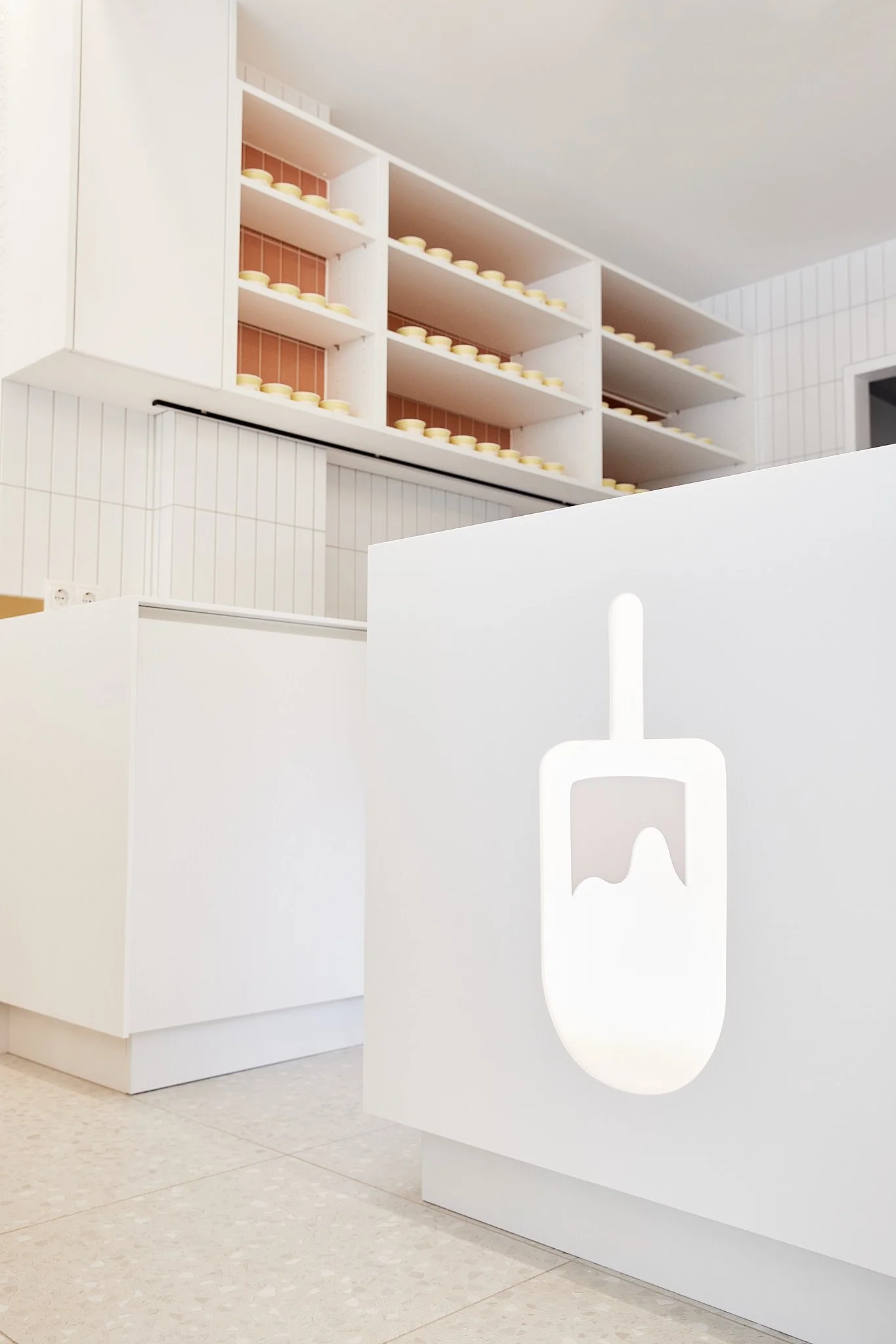 Weißer Tresen mit einem beleuchteten Logo eines Babyfläschchens, im Hintergrund offene Regale mit kleinen Gegenständen, wahrscheinlich Töpfen oder Deckeln, in einem modernen, hellen Raum. Fotografiert von Sarah Neuner.