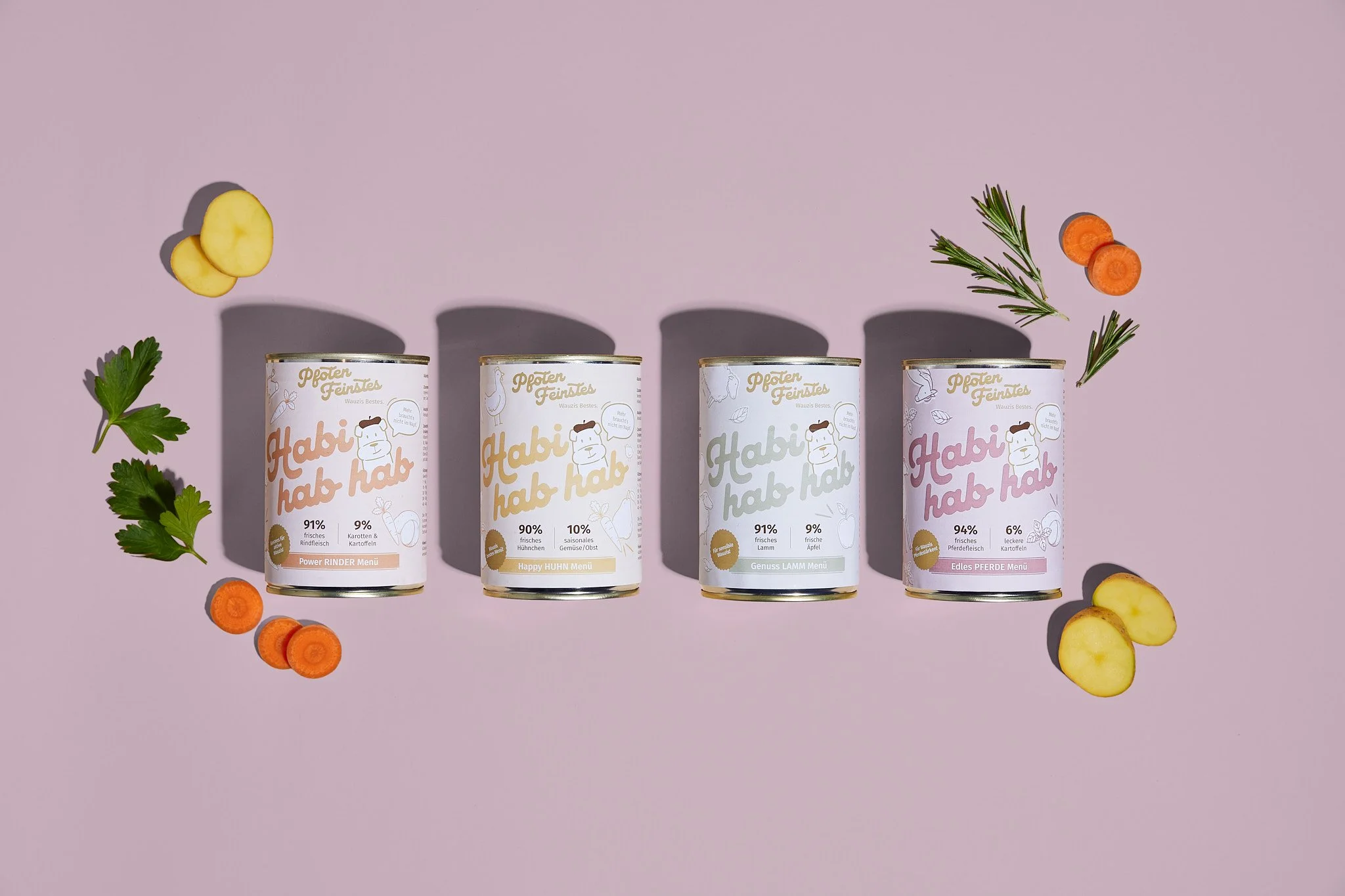 Vier Dosen Haustierfutter mit verschiedenen Geschmacksrichtungen auf rosa Hintergrund, umgeben von Karotten, Kartoffeln, Petersilie und Rosmarin. Fotografiert von Sarah Neuner.
