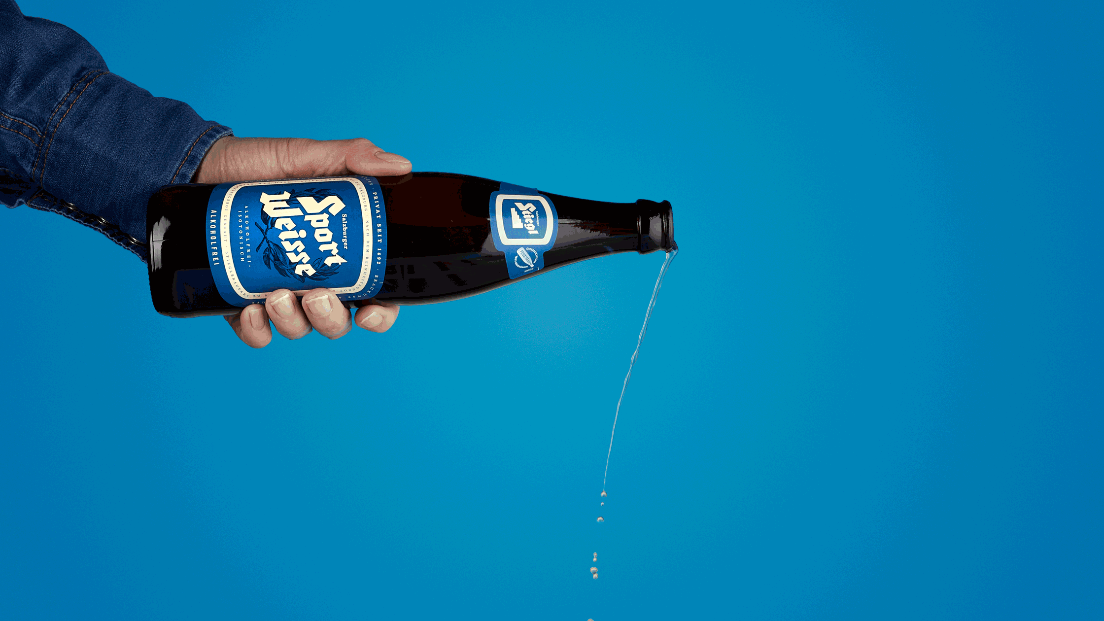 Ein Arm in einer blauen Jacke hält eine dunkle Flasche Bier gegen den blauen Hintergrund, die Flüssigkeit wird in kleinen Tropfen entlassen. Fotografiert von Sarah Neuner.