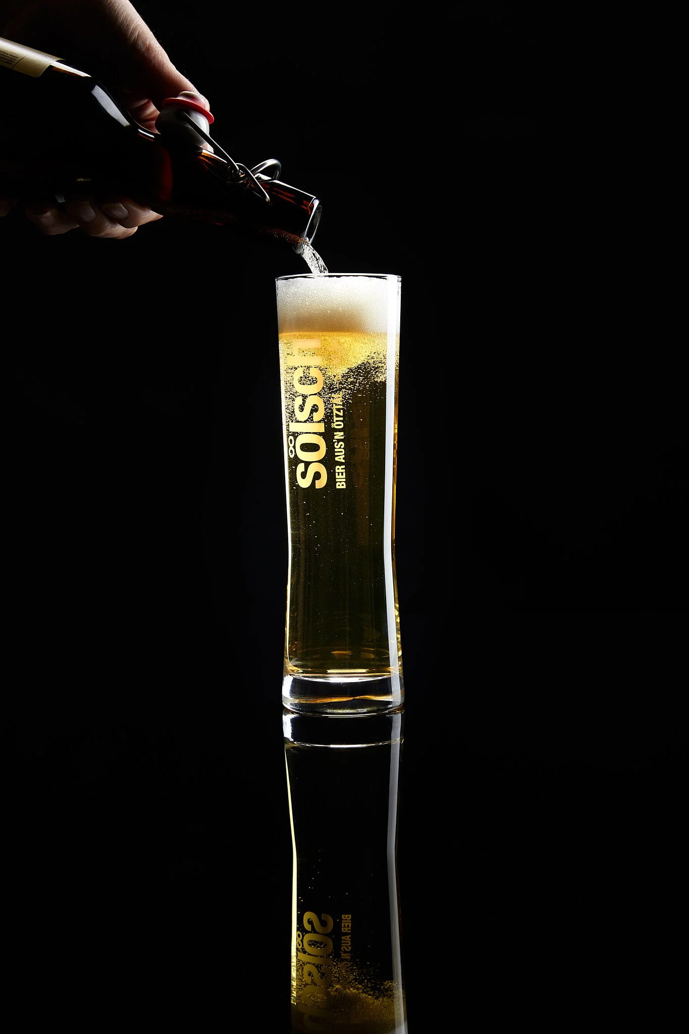 Ein Hand gießt Bier aus einer Flasche in ein hohes, schmales Bierglas, das auf einem schwarzen Hintergrund steht. Fotografiert von Sarah Neuner.