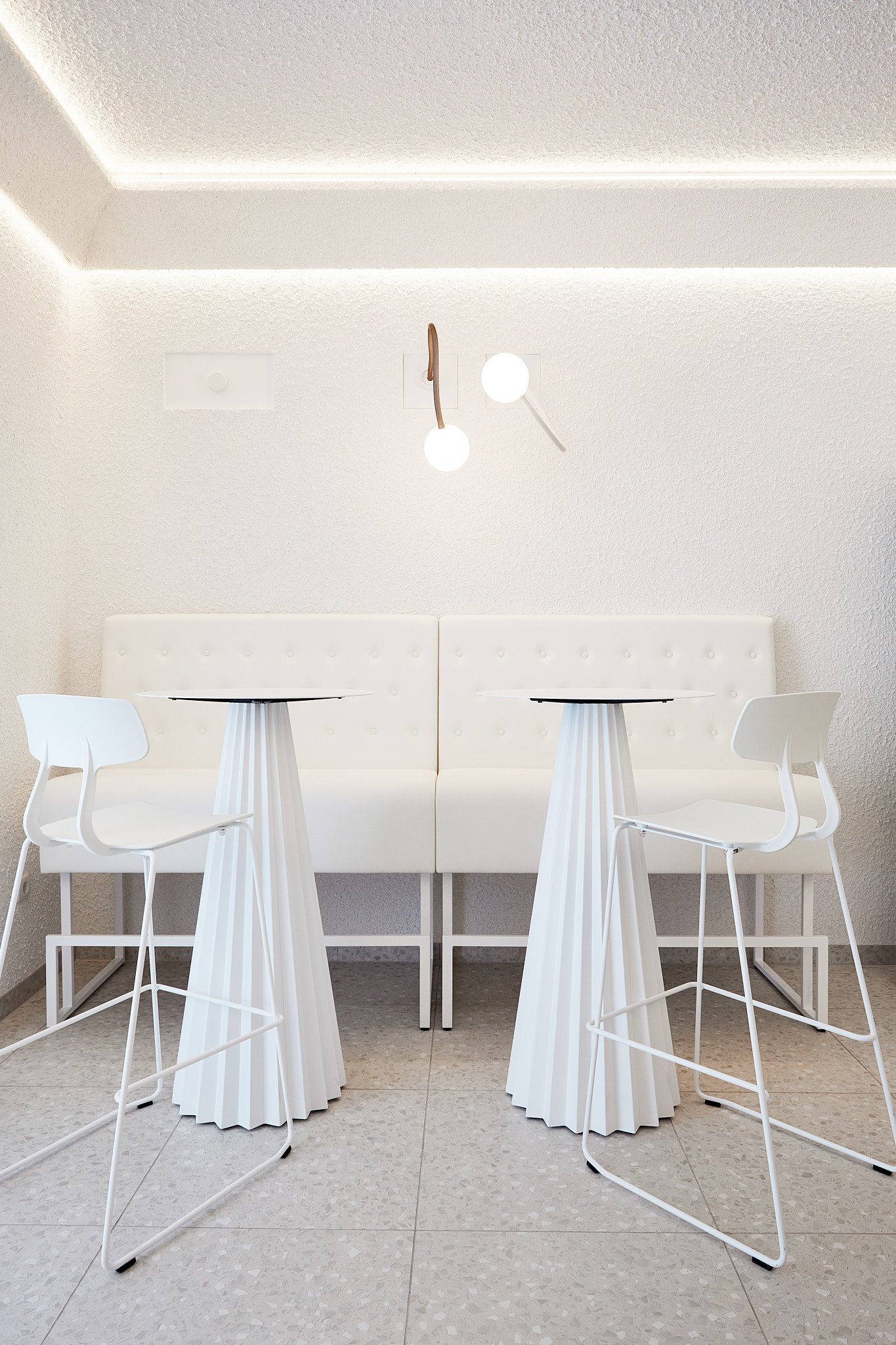 Weiße moderne Sitzecke mit zwei hohen Hockern, kleinen Tischen, weißem Polsterbat, an der Wand mit Beleuchtung. Die Einrichtung ist minimalistisch im skandinavischen Stil. Fotografiert von Sarah Neuner.