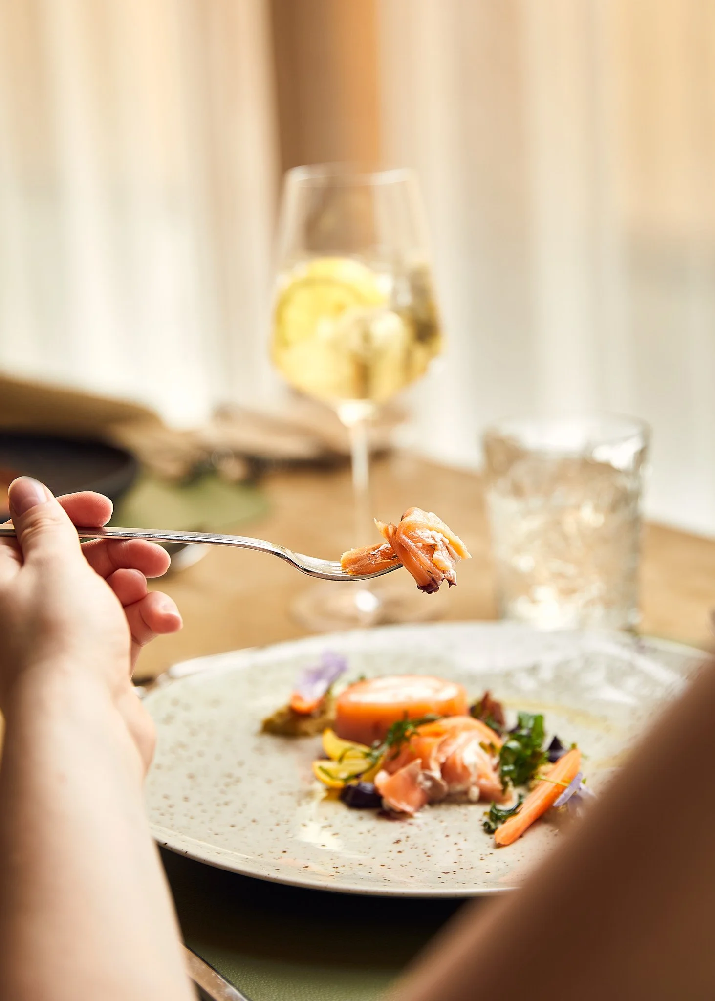 Person isst Fisch mit Gemüse auf einem Teller, im Hintergrund ein Glas Wasser und ein Weinglas mit Zitronenscheibe. Fotografiert von Sarah Neuner.