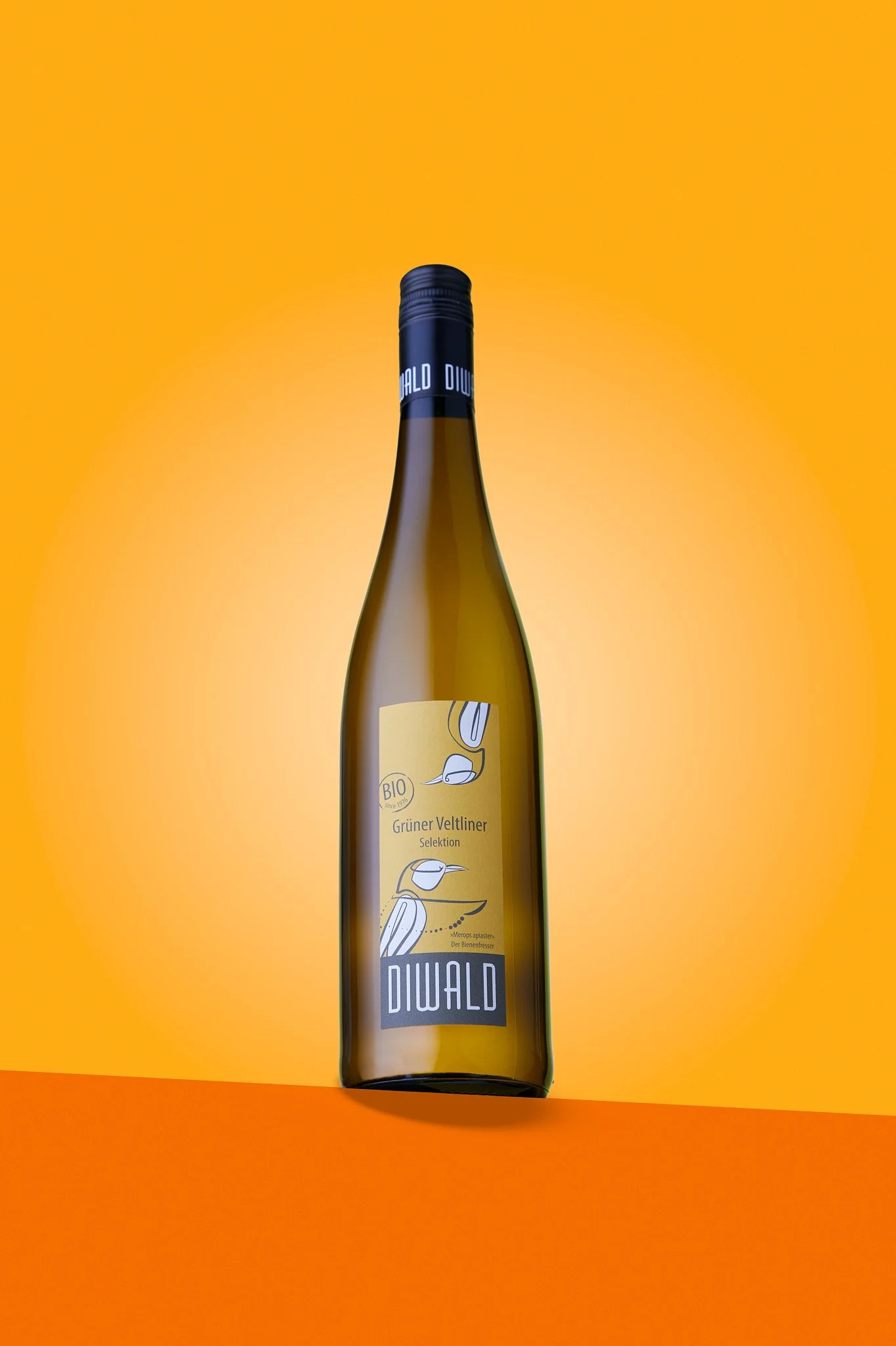 Eine Flasche Weißwein mit gelbem Etikett und blauer Kappe vor einem orangefarbenen Hintergrund. Fotografiert von Sarah Neuner.