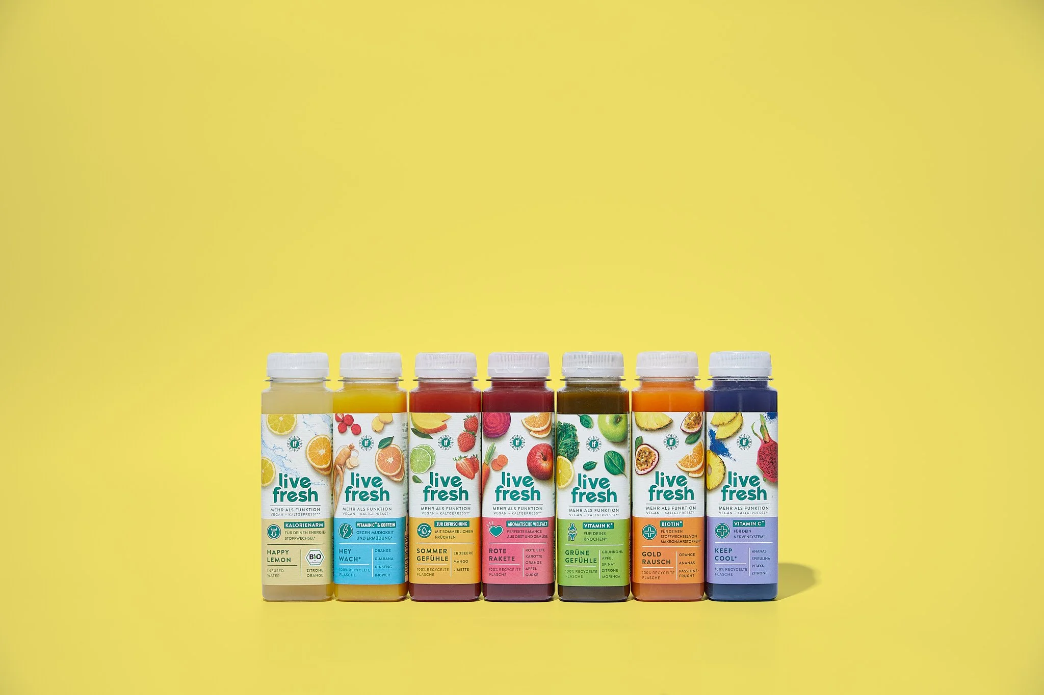 Sechs Flaschen mit verschiedenen Frucht-Getränken auf gelbem Hintergrund, jede Flasche zeigt Früchte und Beschreibungen auf dem Etikett. Fotografiert von Sarah Neuner.