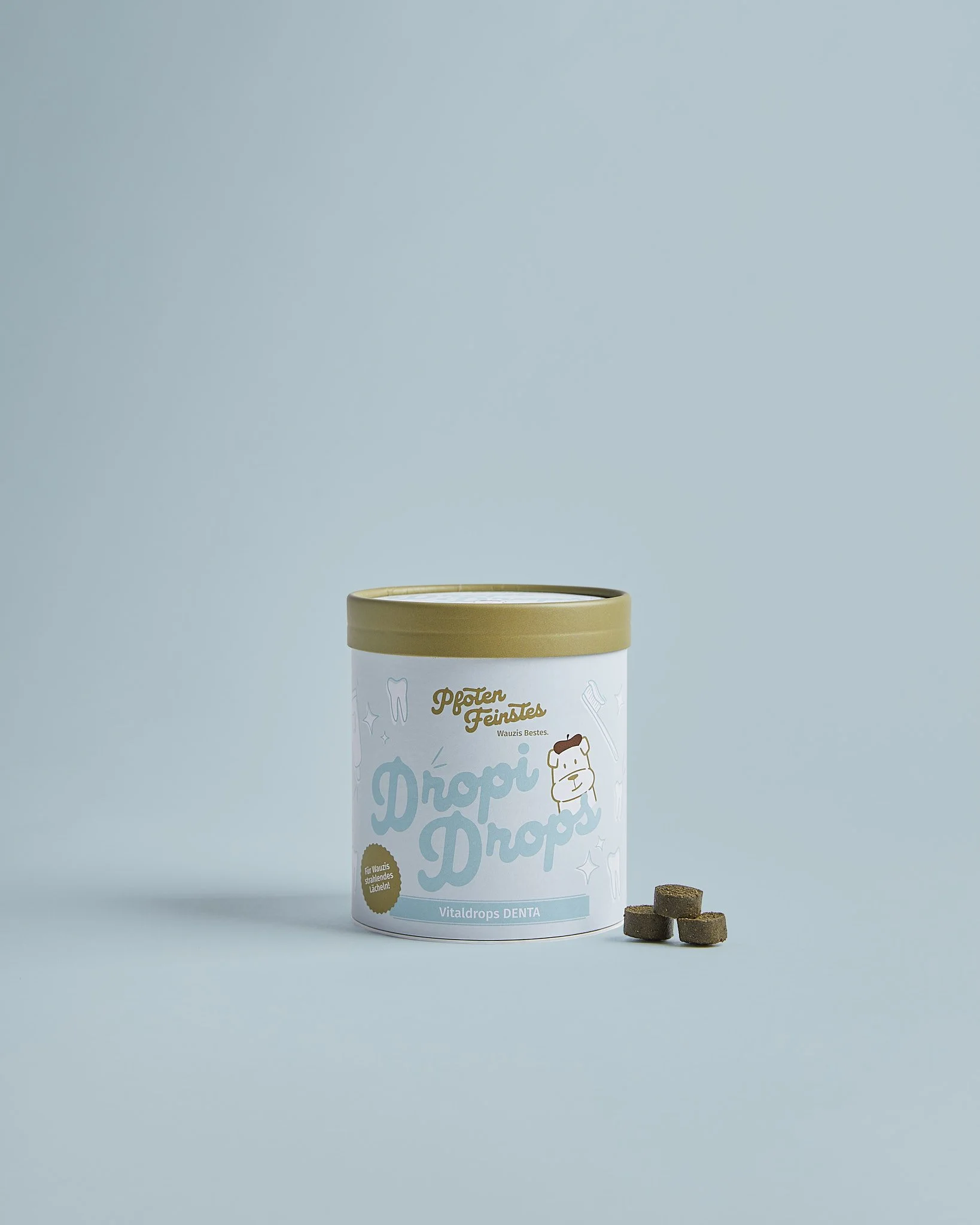 Ein runder Blechdosenbehälter mit gelbem Deckel, der auf einem hellblauen Hintergrund steht. Auf der Dose befindet sich eine Illustration eines Hundes und der Text "Dropi Drops" sowie zusätzliche Beschriftungen und Designs. Neben der Dose liegen vier