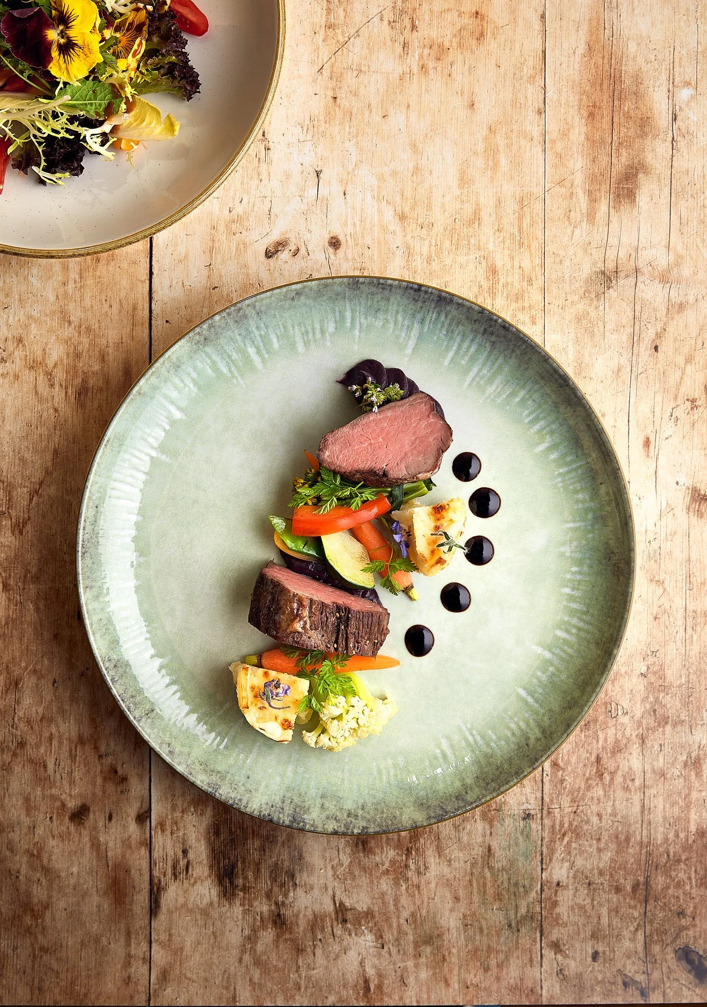 Auf einem grünen Teller ist ein Gericht mit verschiedenen Fleischstücken, Gemüse und schwarzen Sauce-Dots. Zusätzlich befindet sich eine Salatschüssel mit grünem und rotem Salat oben links auf dem Bild.