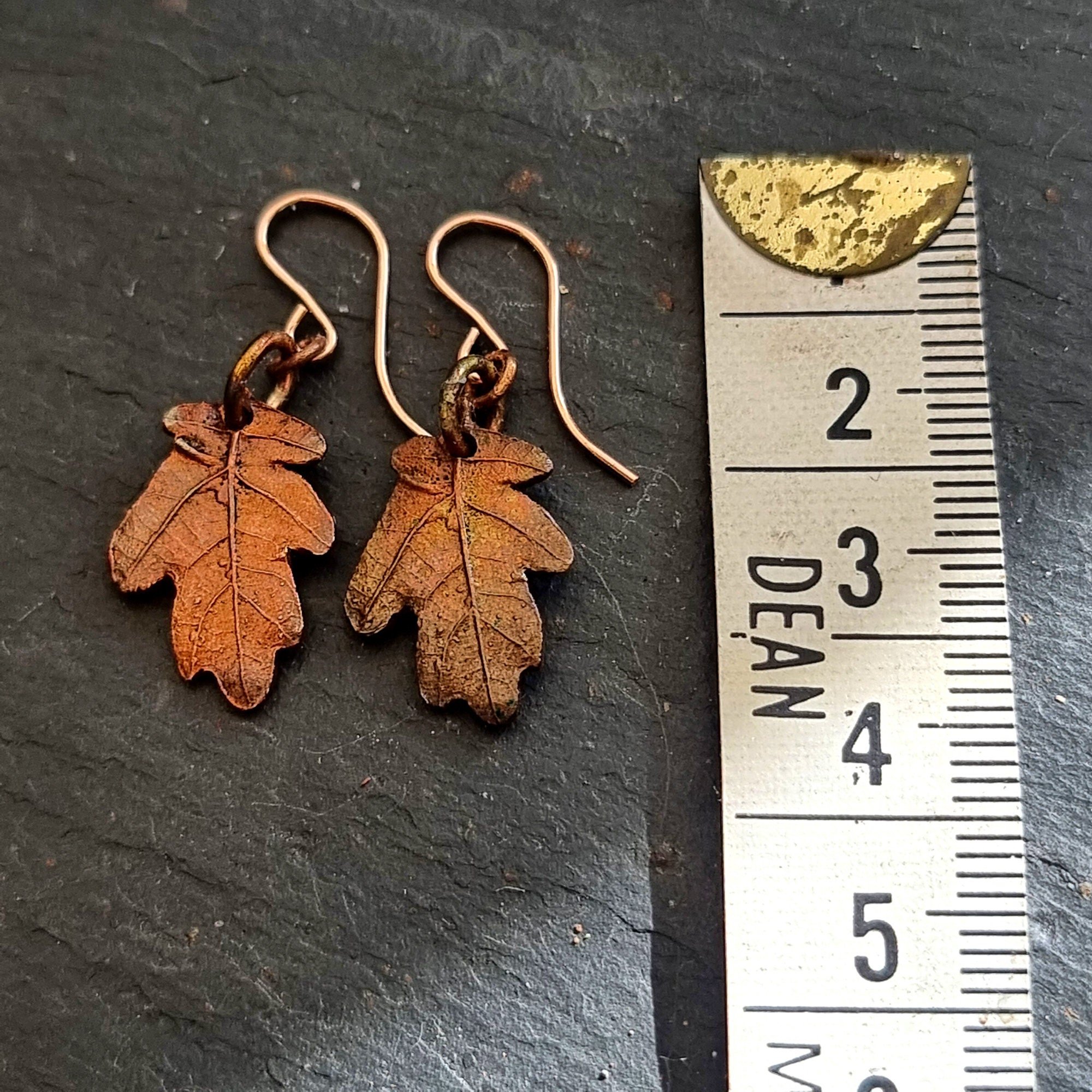 Two copper oak leaf earrings lying next to a measuring tape showing centimetres on a dark slate background.