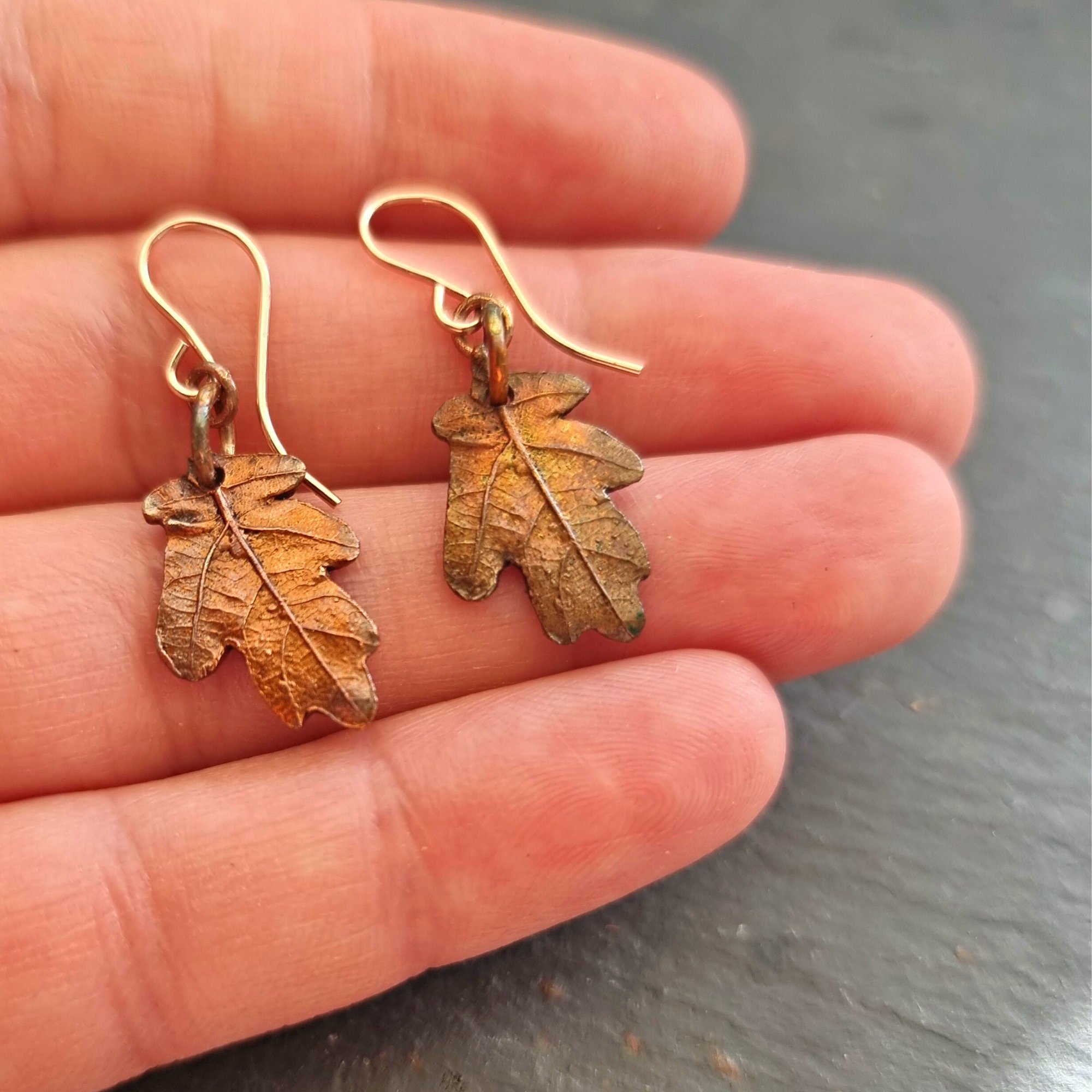 Two copper oak leaf earrings on rose gold ear hooks lying on a hand to show scale of earrings.